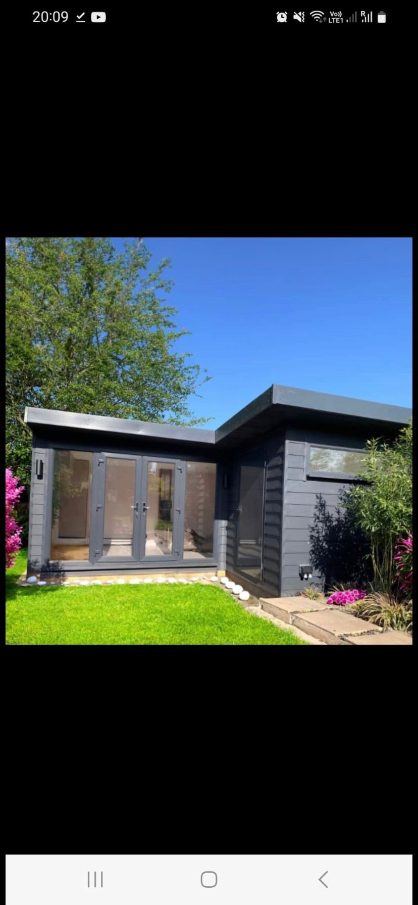 Nowoczesny, szary domek ogrodowy z dużymi oknami i drzwiami, otoczony zielonym trawnikiem i kwitnącymi krzewami w słoneczny dzień.