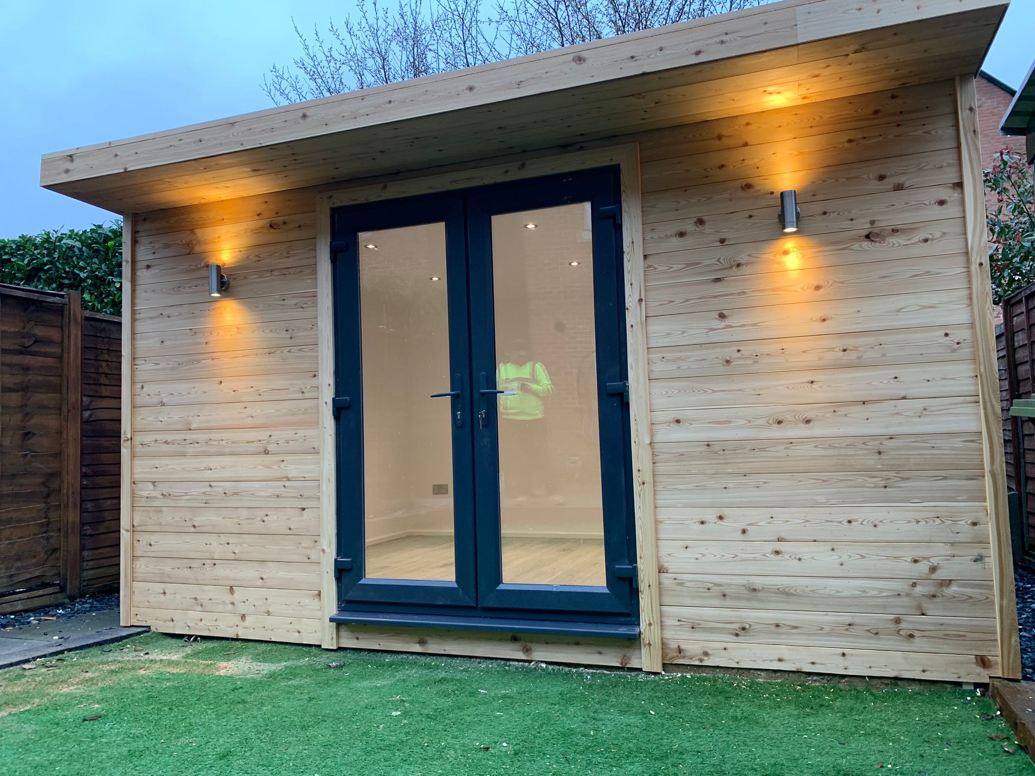 Nowoczesny domek ogrodowy z drewnianą elewacją, oświetleniem zewnętrznym i dwuskrzydłowymi drzwiami w kolorze antracytowym, widok z zewnątrz na sztuczną trawę.
