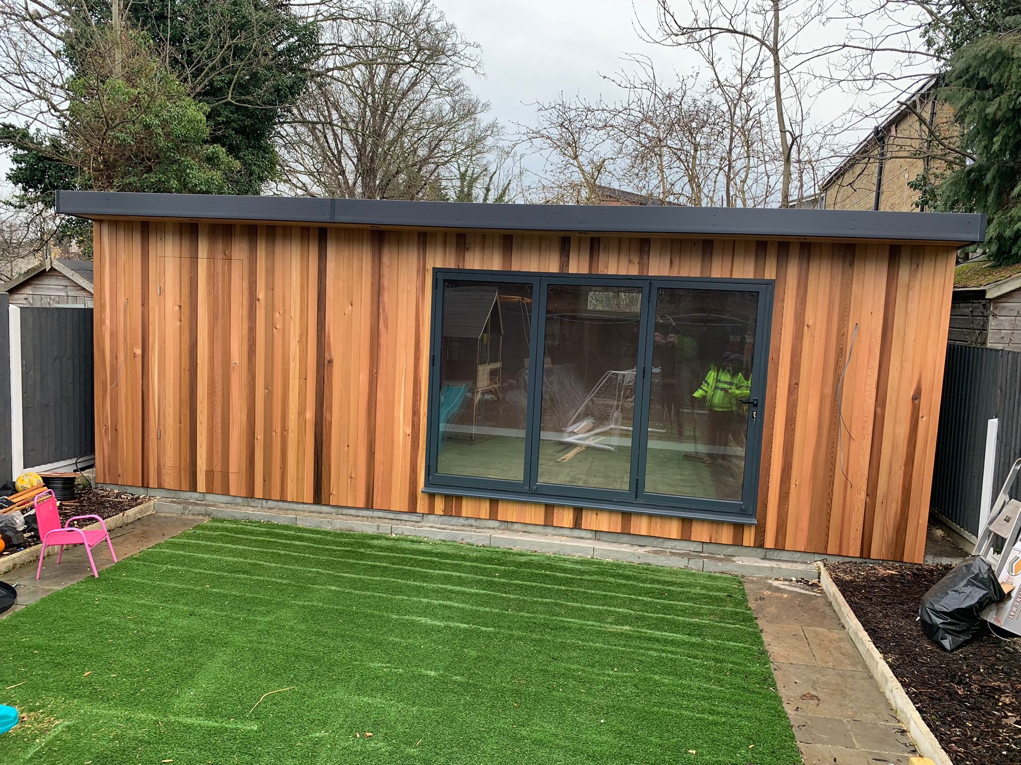 Nowoczesny, drewniany domek ogrodowy z dużymi, składanymi drzwiami szklanymi, otoczony sztuczną trawą i ogrodzeniem, z widocznymi drzewami w tle.