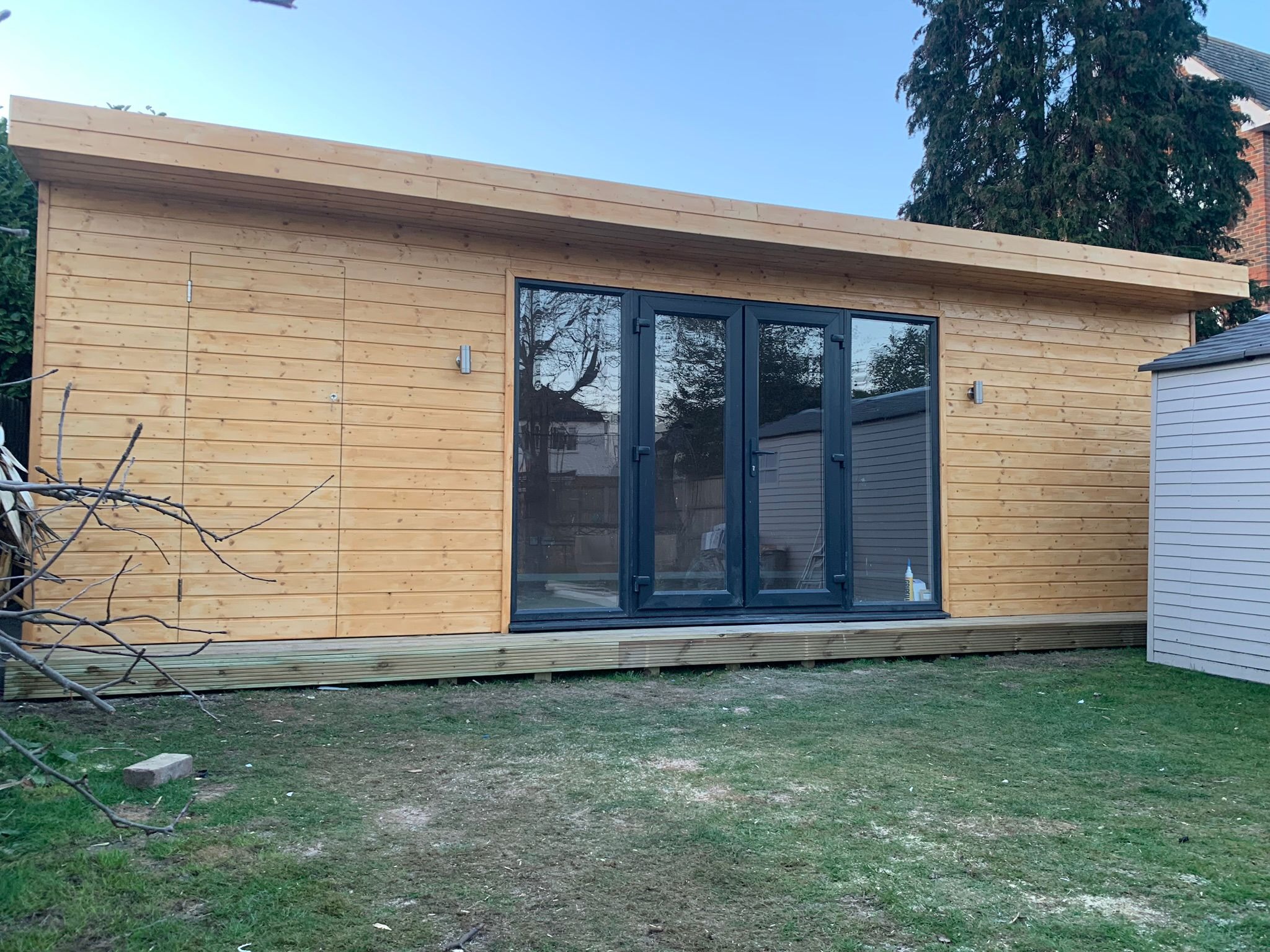 Nowoczesny, drewniany domek ogrodowy z ciemnymi, przeszklonymi drzwiami, otoczony trawnikiem. Widoczne elementy oświetlenia zewnętrznego.