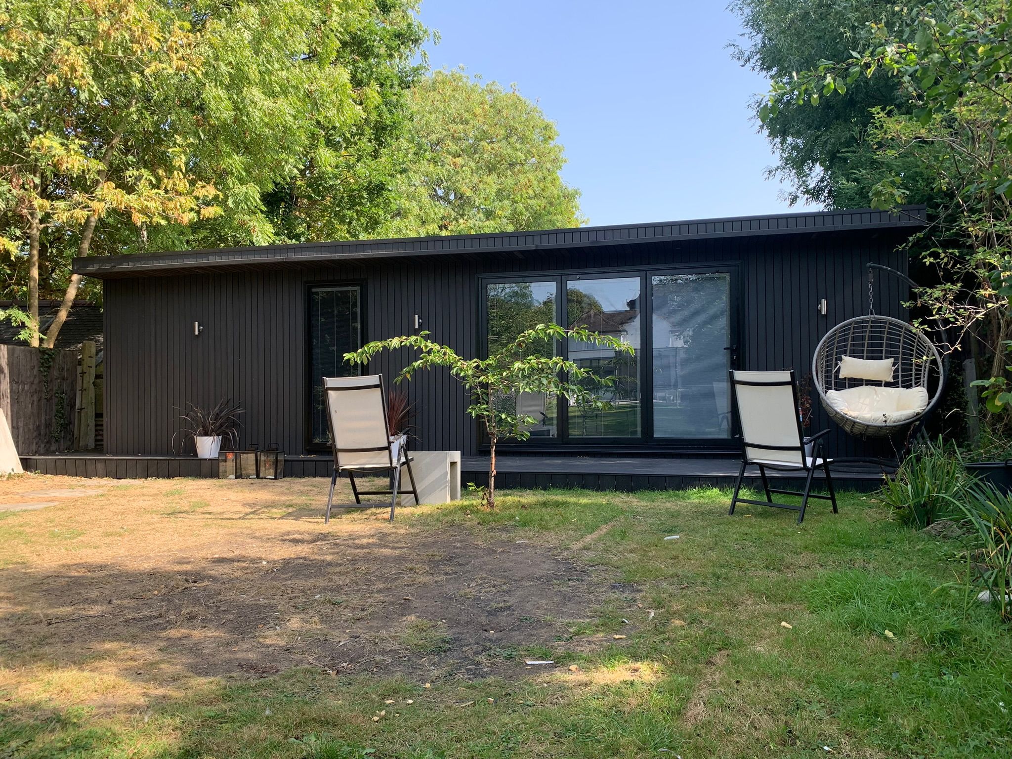 Nowoczesny, ciemny domek letniskowy z dużymi oknami przesuwnymi, otoczony zielenią, z wiszącym fotelem i krzesłami na zewnątrz, z widocznym fragmentem trawnika.