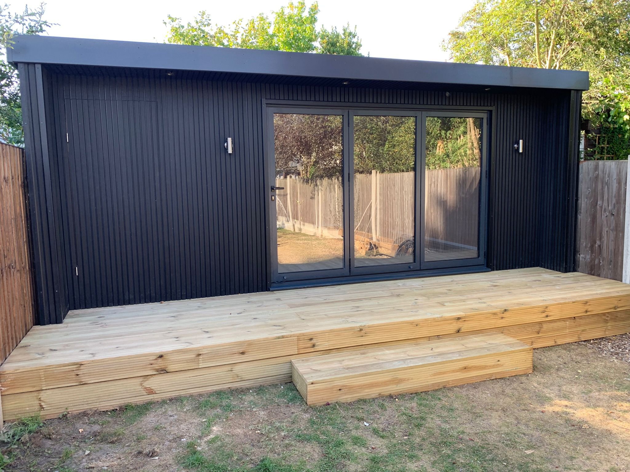 Nowoczesna, czarna altana z przesuwnymi szklanymi drzwiami i drewnianym tarasem z dwoma stopniami, na trawniku.