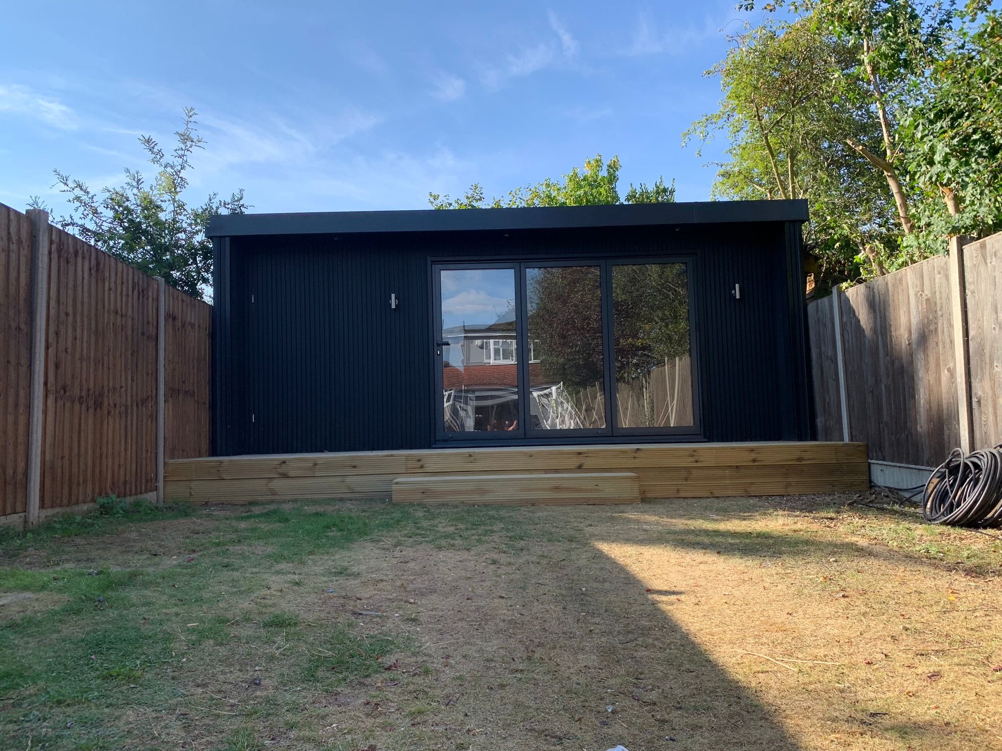 Nowoczesny, czarny domek ogrodowy z przesuwnymi drzwiami i drewnianym podestem, otoczony trawnikiem i drewnianym ogrodzeniem.