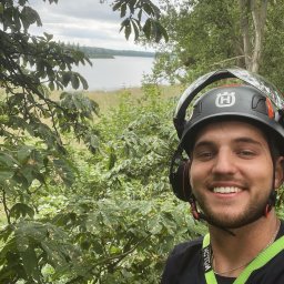 TedTree - Uśmiechnięty mężczyzna w kasku ochronnym Husqvarna, z słuchawkami i uprzężą, pozuje na tle zieleni i jeziora podczas pracy.