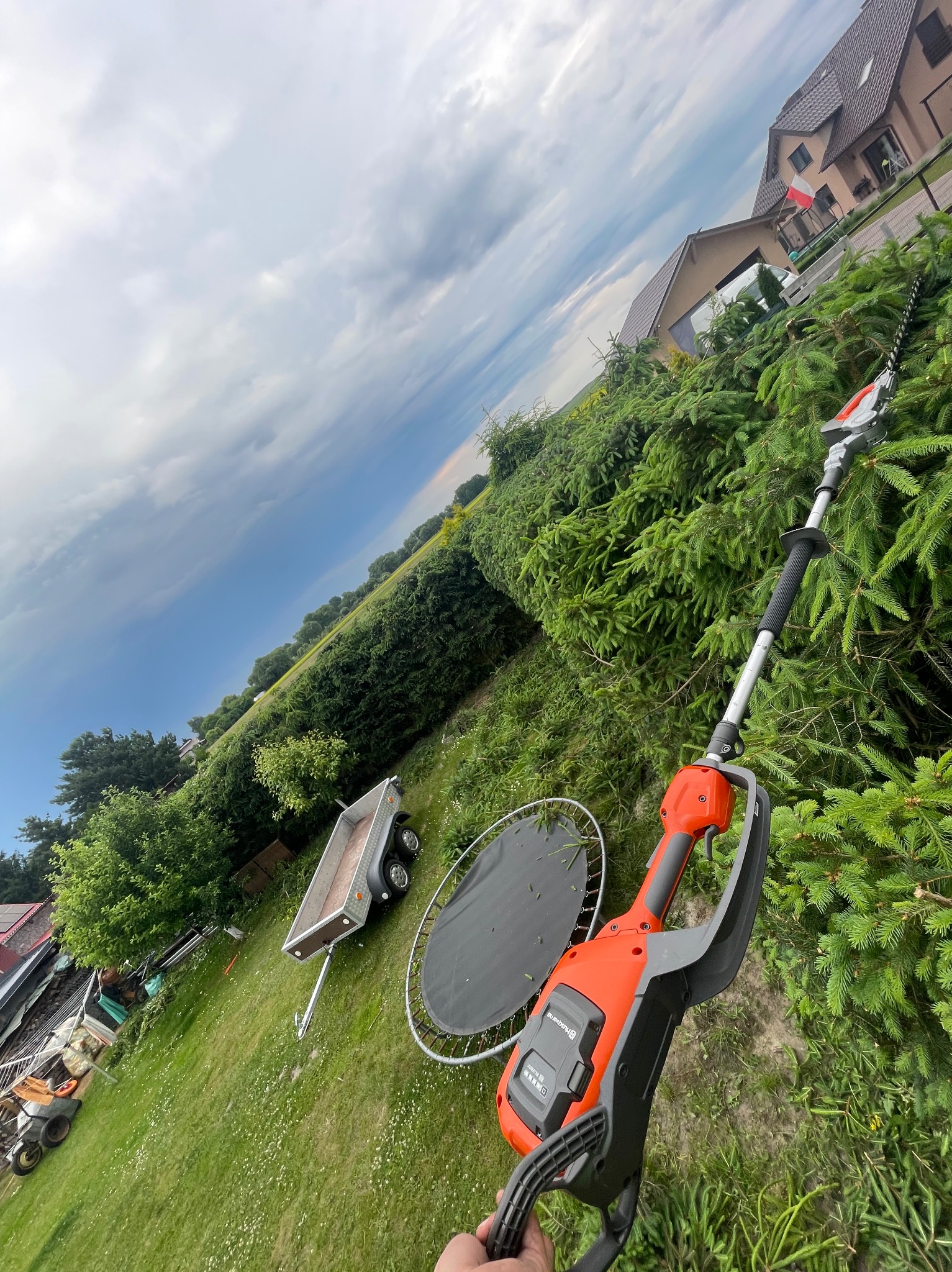 Elektryczne nożyce do żywopłotu na wysięgniku, z przyczepką i trampoliną w tle, podczas formowania żywopłotu z iglaków w ogrodzie.