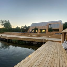 Skandi Pur - Nowoczesny dom z drewnianą elewacją i rozległym tarasem nad wodą oświetlonym punktowymi lampami, widok na zachód słońca.