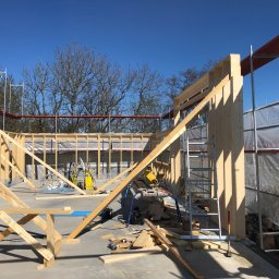 Skandi Pur - Konstrukcja szkieletowa domu w trakcie budowy, widoczne drewniane elementy, rusztowania i narzędzia pracy, jasne światło słoneczne.