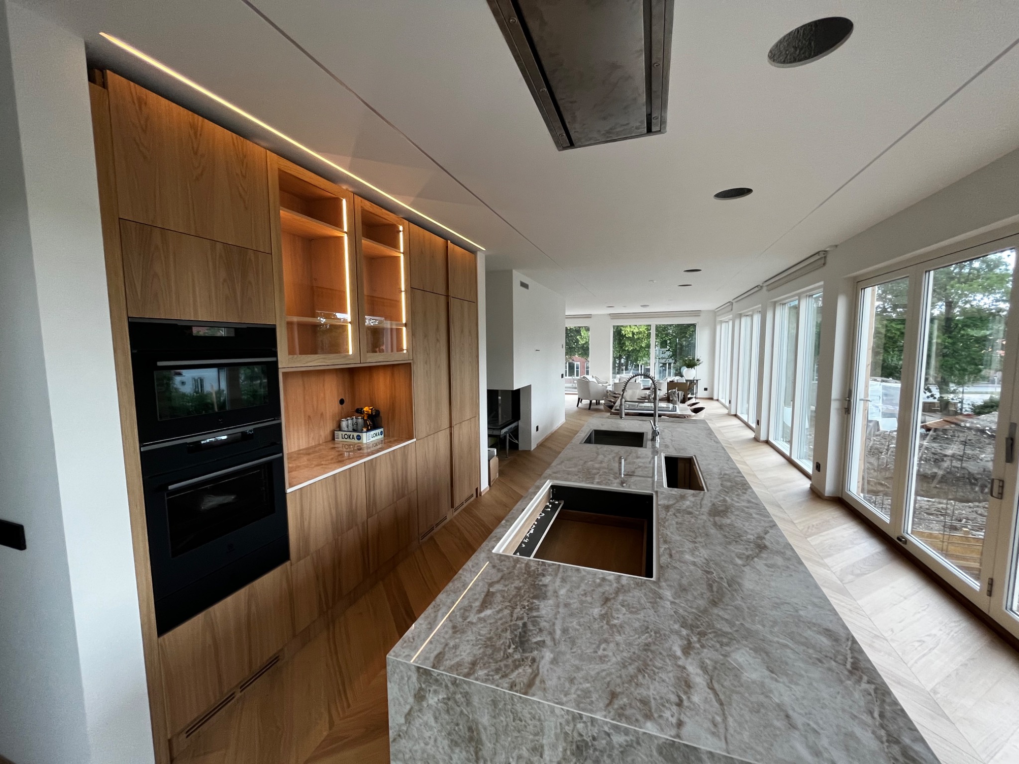 Nowoczesna kuchnia z drewnianą zabudową, marmurowym blatem wyspowym z dwoma zlewozmywakami, oświetleniem LED podszafkowym i wnękowym oraz widokiem na ogród przez duże okna.