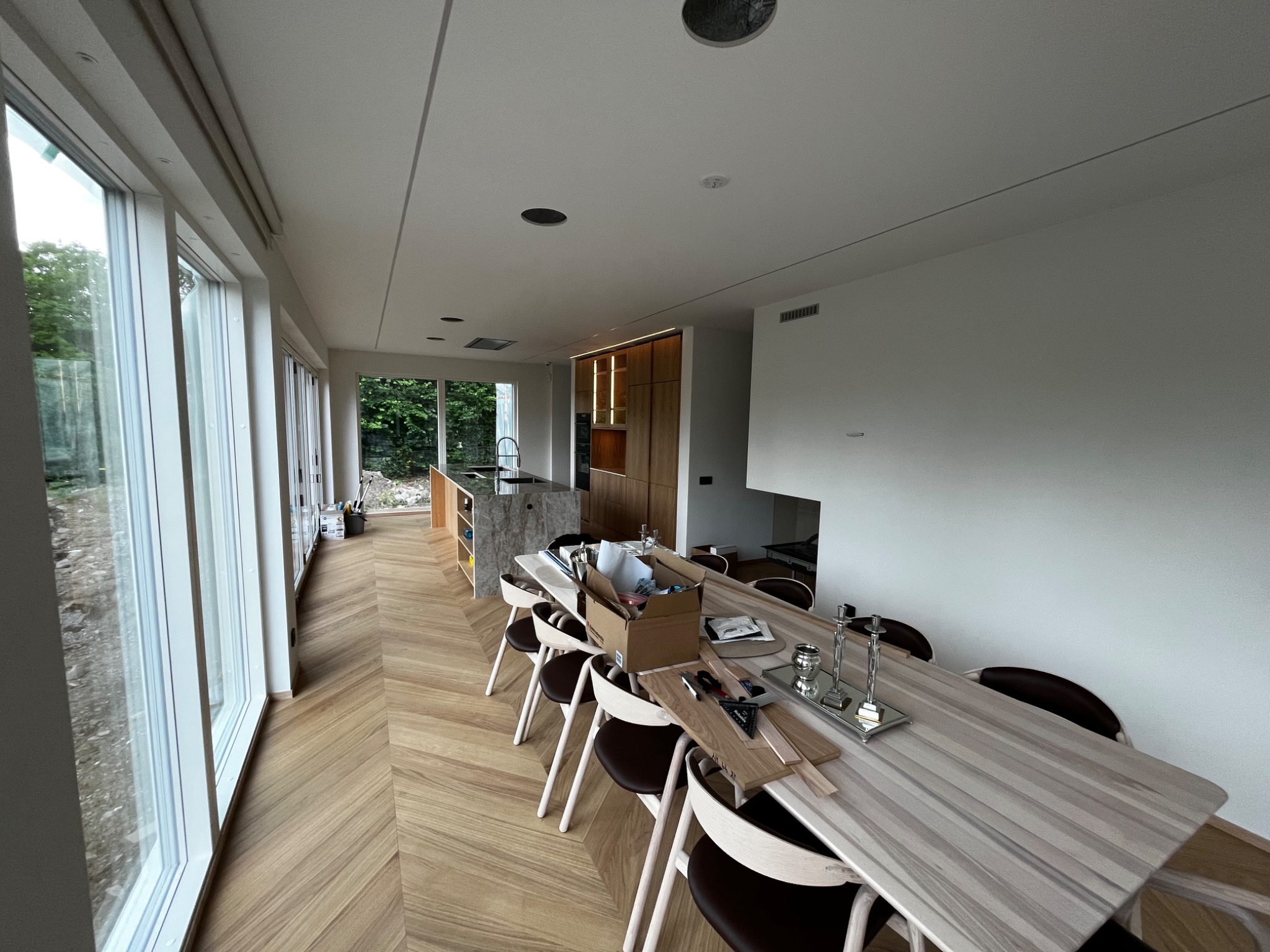 Wnętrze domu w trakcie wykończenia: parkiet ułożony w jodełkę, duży stół z krzesłami, narzędzia i materiały budowlane na blacie, widok na kuchnię i ogród przez duże okna.