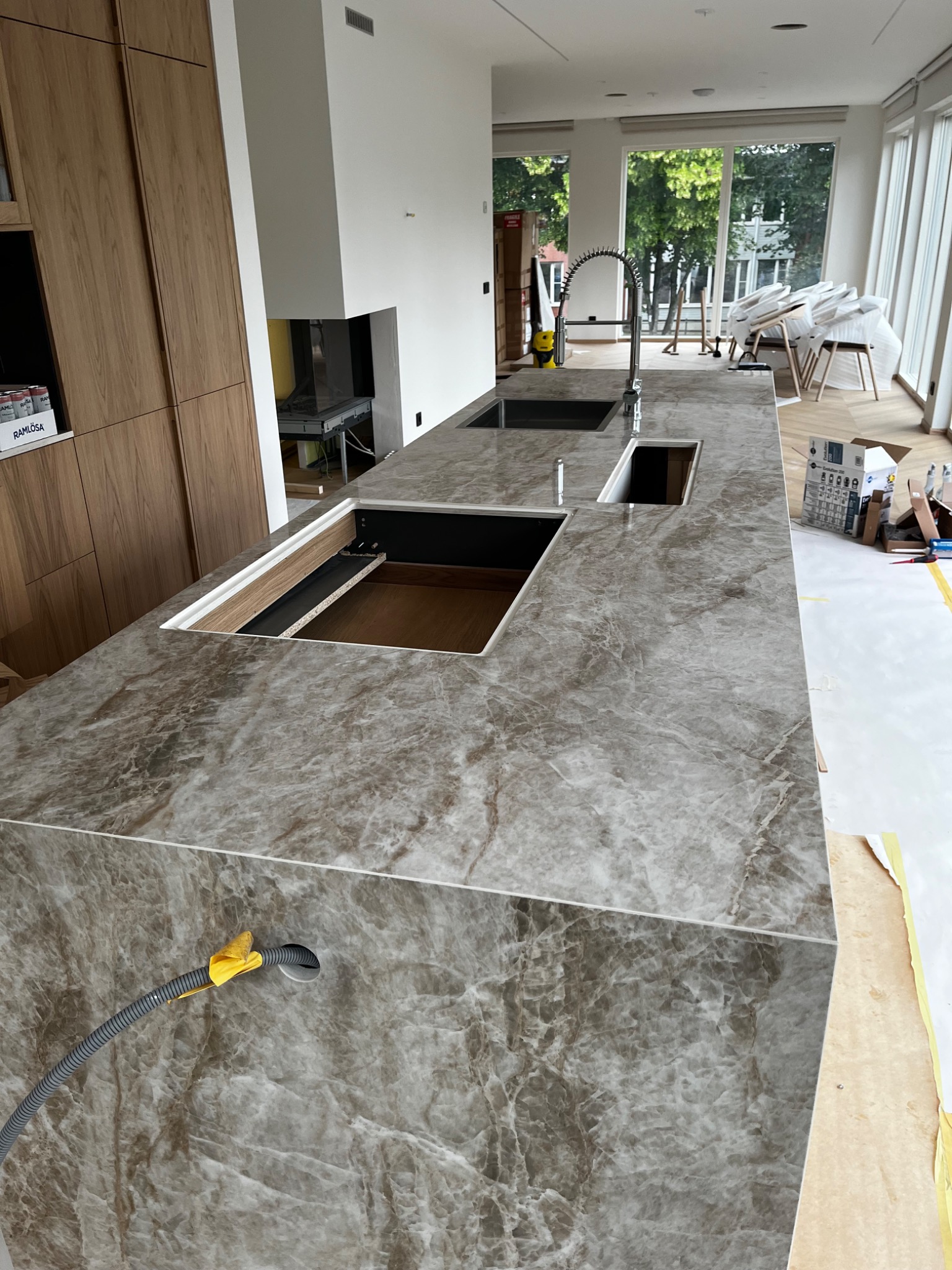 Montaż blatu kuchennego z szarego marmuru z wyciętymi otworami na zlew i płytę. Widoczne zabezpieczenia podłogi i narzędzia.