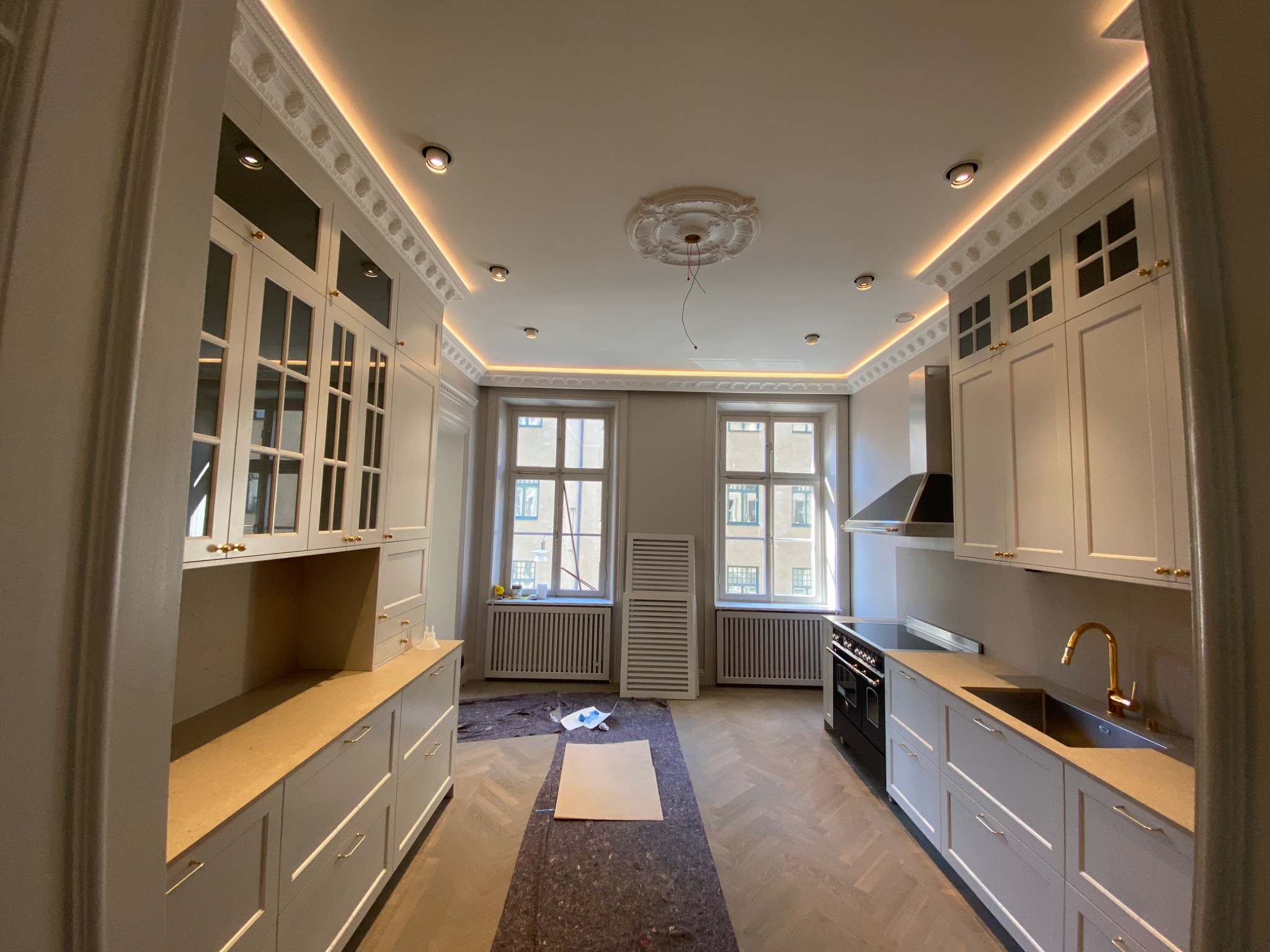 Jasna kuchnia w klasycznym stylu, z białymi szafkami z przeszklonymi górnymi drzwiczkami, złotymi uchwytami i podświetlanym sufitem z dekoracyjną sztukaterią, w trakcie prac wykończeniowych...