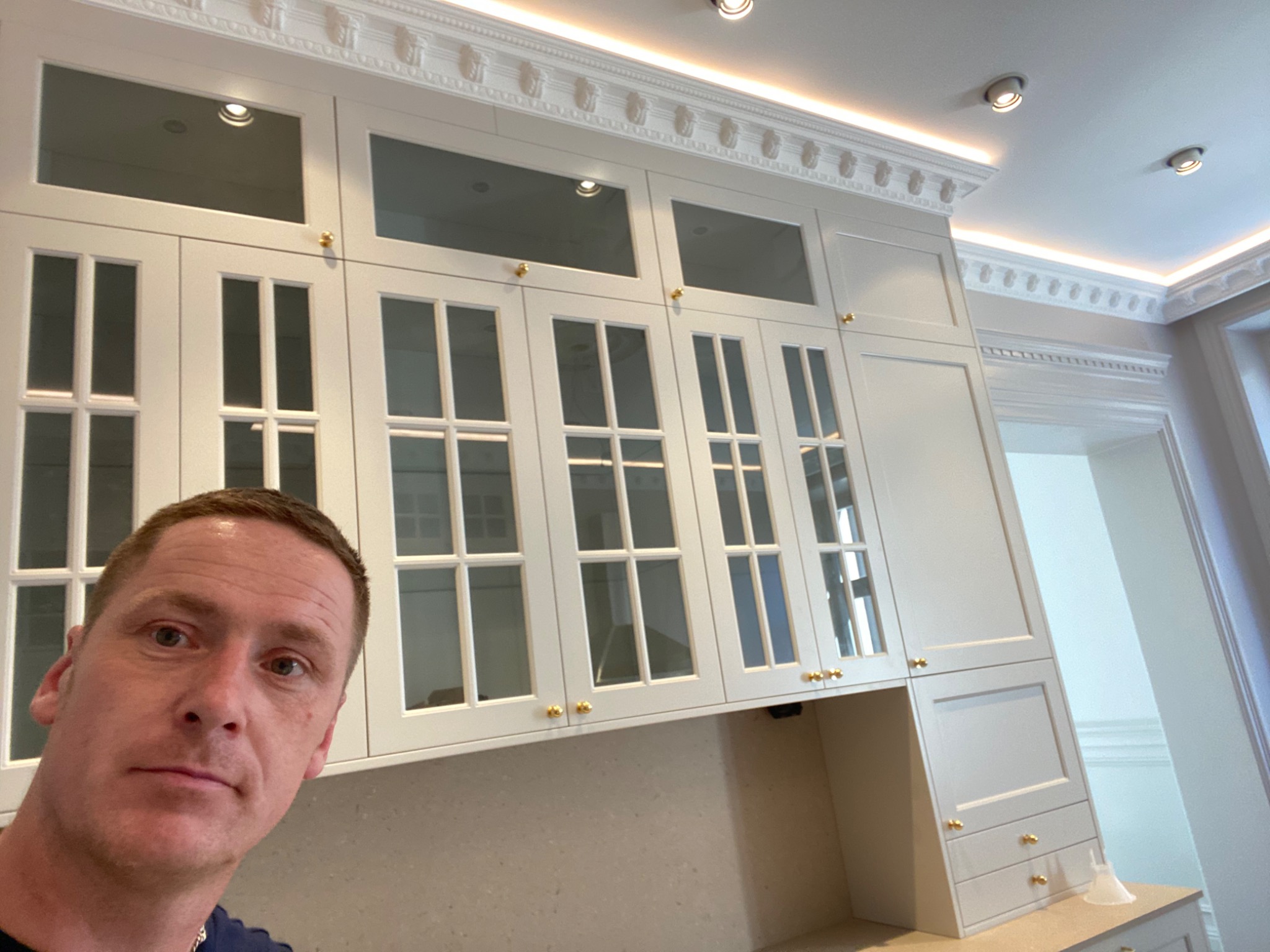 Człowiek robiący selfie na tle białych, przeszklonych szafek kuchennych z dekoracyjnymi listwami przysufitowymi i złotymi gałkami, widoczne oświetlenie punktowe.