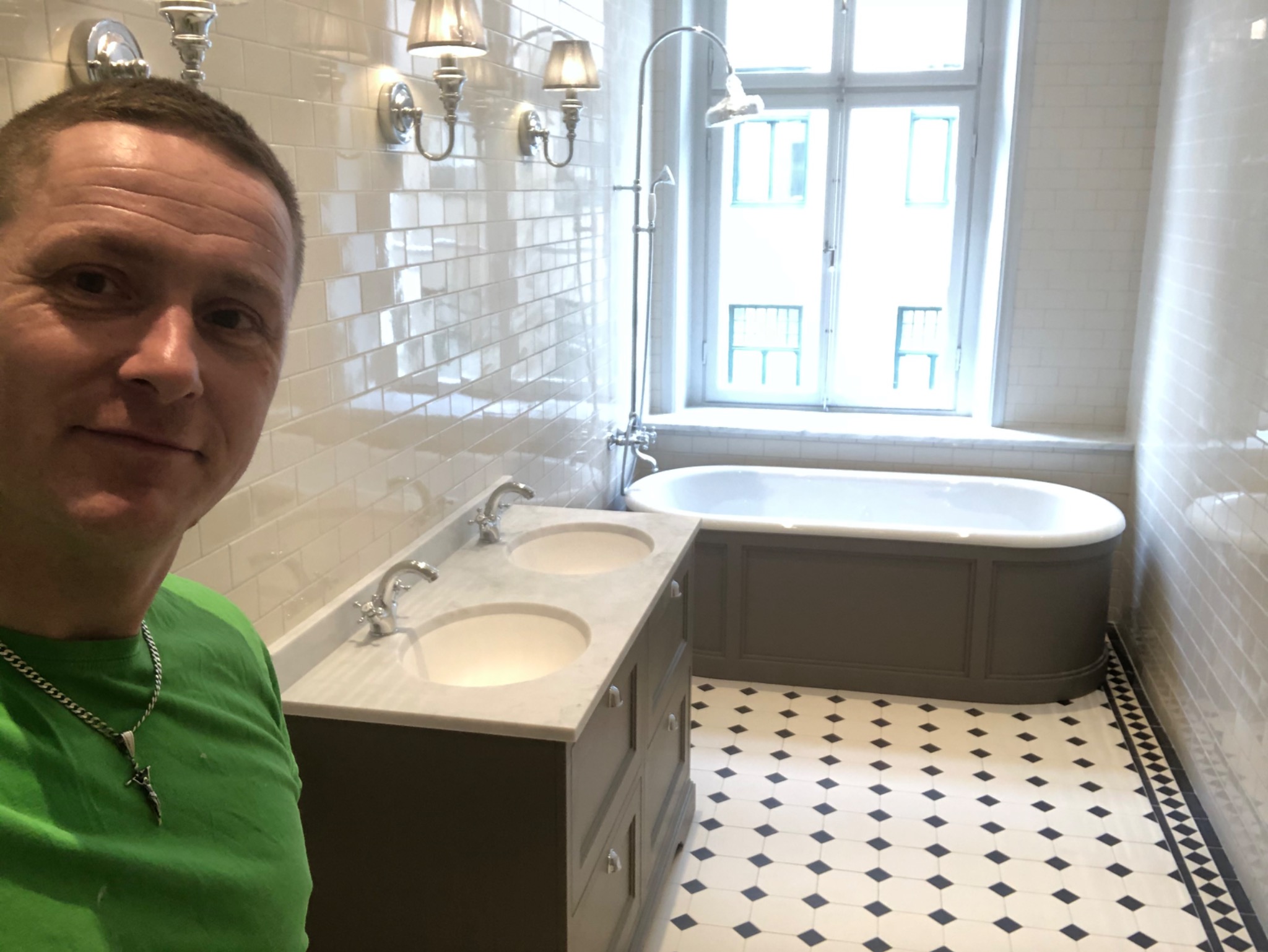 Wykończona łazienka w Kolbudach z wanną na tle okna i umywalką z szafką, białe płytki na ścianach, czarno-biała podłoga w karo, widoczny pracownik w zielonej koszulce.