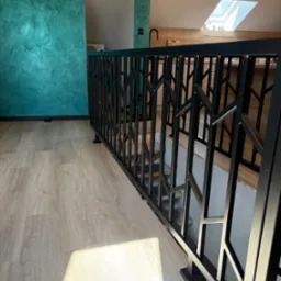 Czarna, metalowa balustrada schodowa o geometrycznym wzorze w nowoczesnym wnętrzu. Widok na schody i turkusową ścianę z przesuwnymi drzwiami.