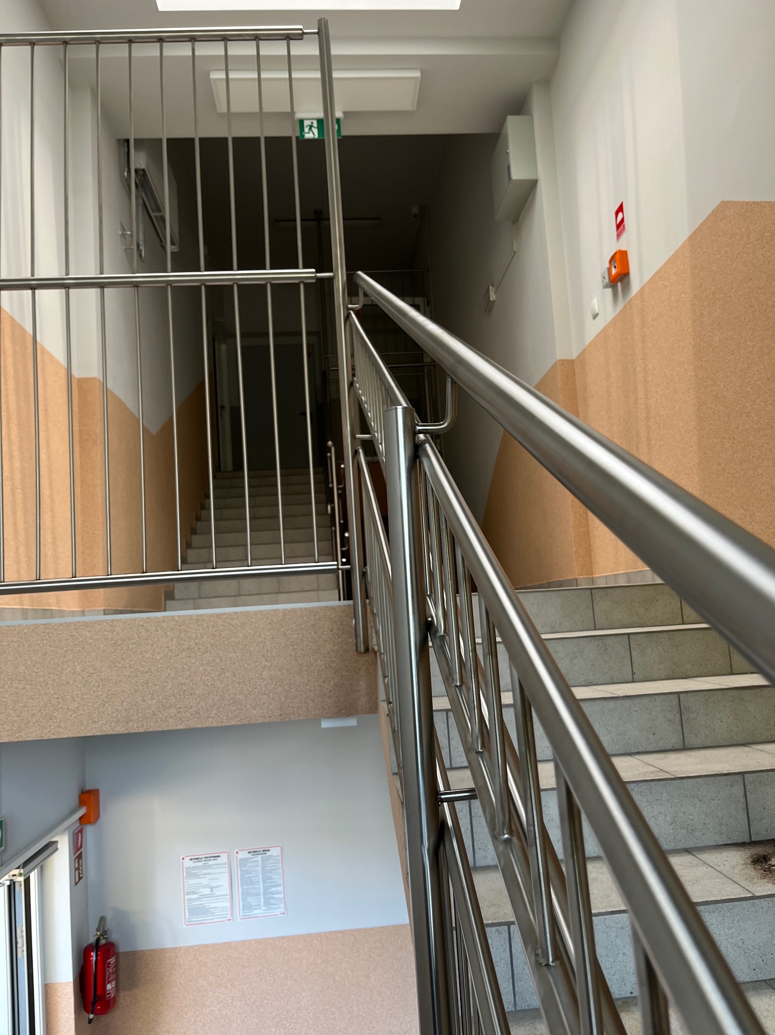 Wewnętrzne schody z metalową balustradą i metalowym zabezpieczeniem na półpiętrze, widok z dołu na górę, na ścianie instrukcje i gaśnica.