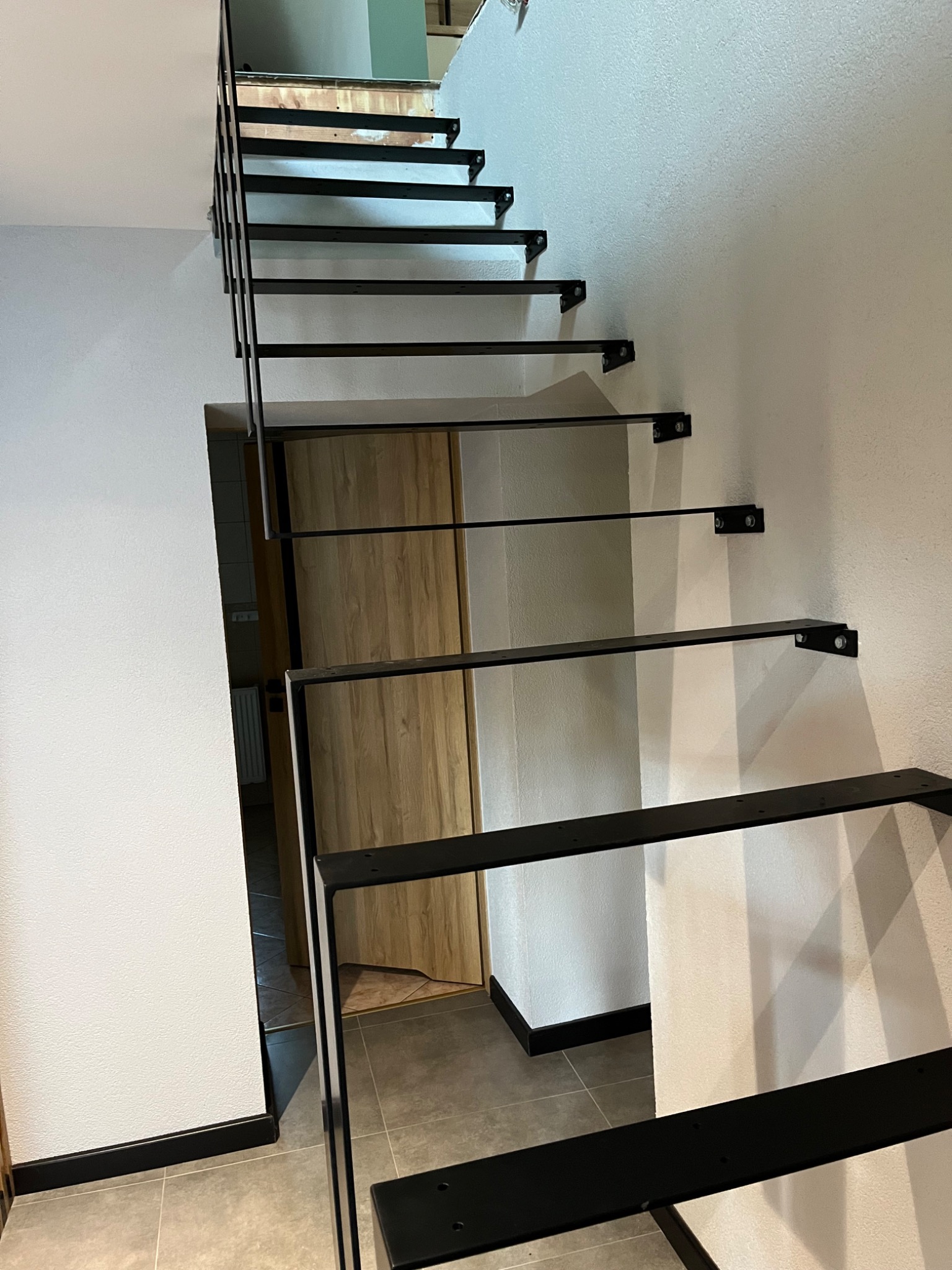 Minimalistyczne schody metalowe z czarnymi, płaskimi stopniami, zamocowane do białej ściany, widok od dołu, drzwi z jasnego drewna w tle.