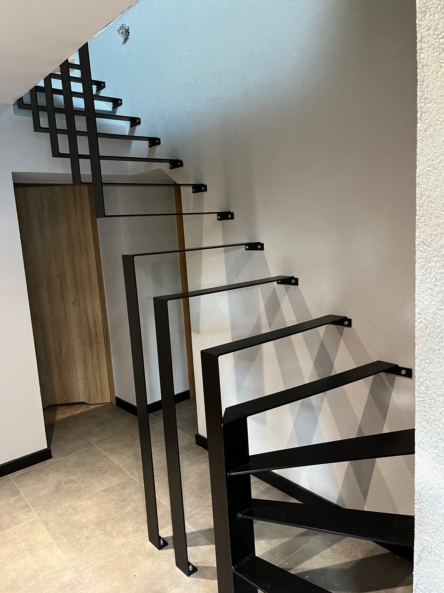 Nowoczesne schody wspornikowe z czarnymi metalowymi stopniami przymocowanymi do ściany, widok z dołu na tle jasnej ściany i drewnianych drzwi.