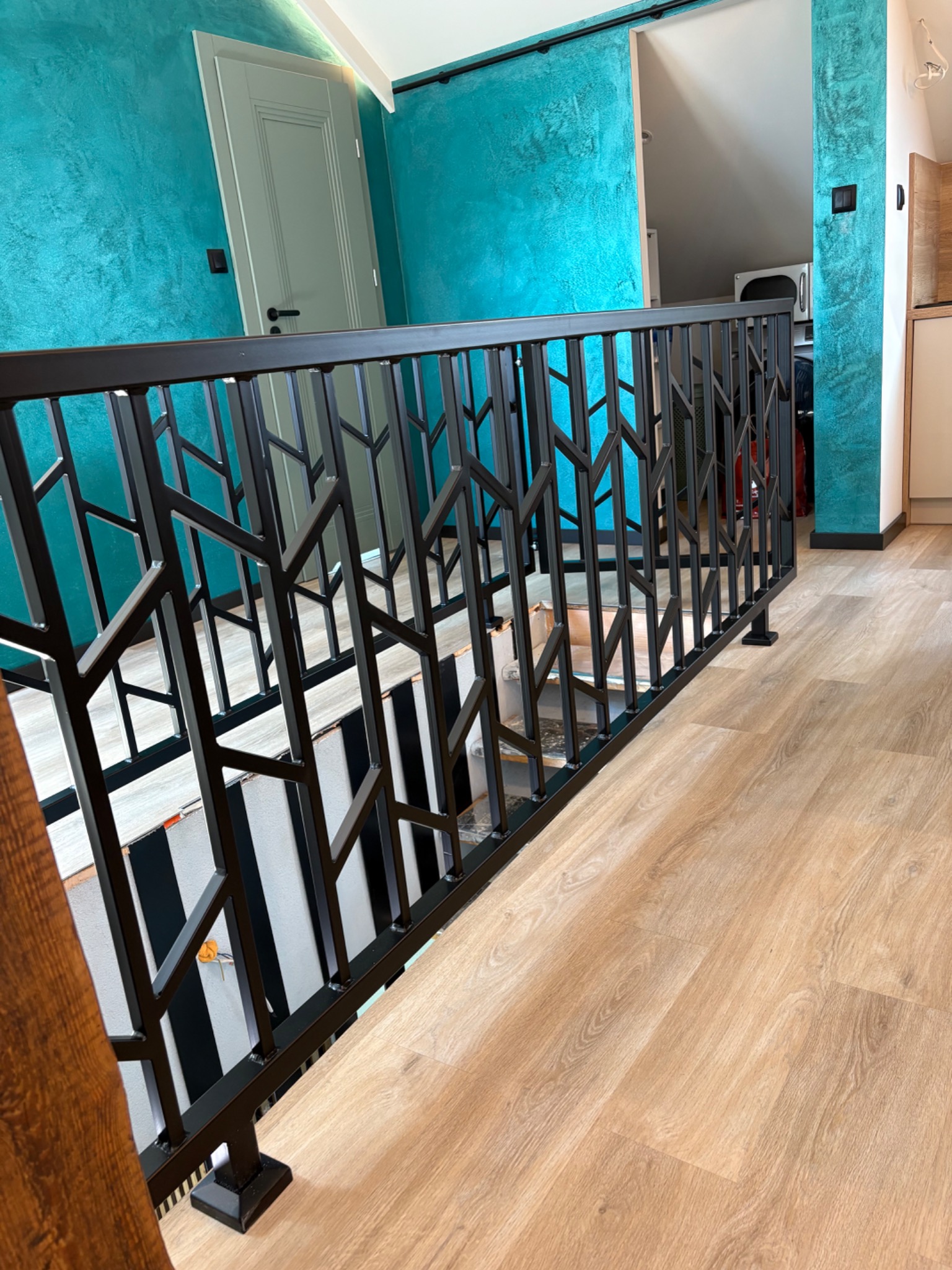 Nowoczesna balustrada schodowa z czarnego metalu o geometrycznym wzorze, na tle turkusowej ściany i jasnej podłogi. Widok z góry na schody.