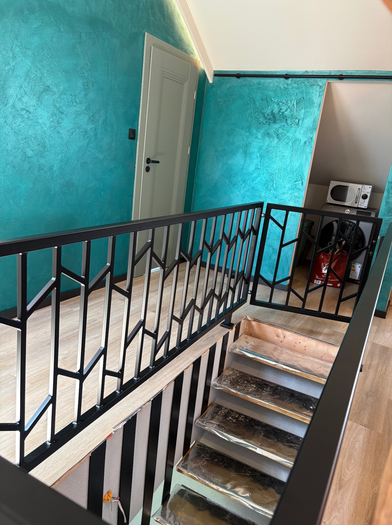 Nowoczesna balustrada schodowa z czarnego metalu o geometrycznym wzorze, zabezpieczająca schody. W tle turkusowa ściana i białe drzwi.
