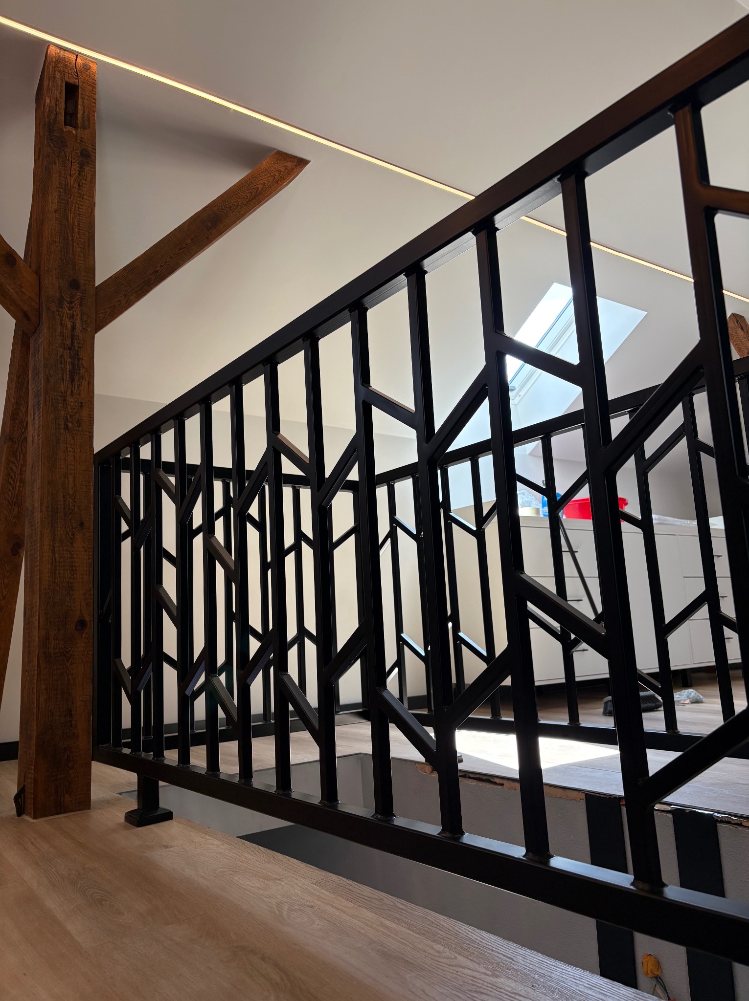 Czarna, metalowa balustrada schodowa z geometrycznym wzorem, drewniany słup wspierający, widok na poddasze z oknem dachowym, nowoczesny design.