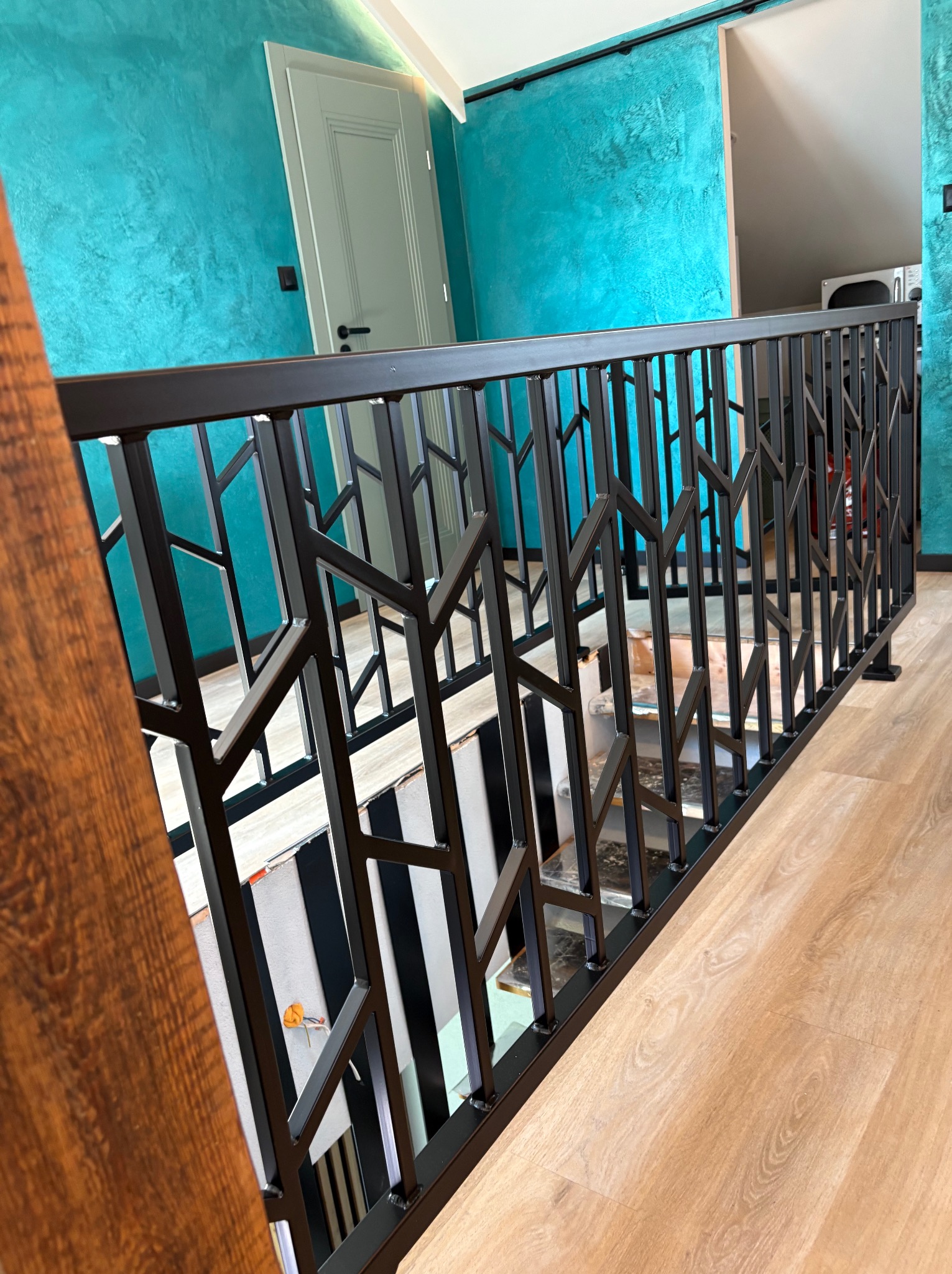 Nowoczesna, czarna balustrada schodowa z geometrycznym wzorem, widok z góry, na tle turkusowej ściany i drewnianej podłogi. Nietypowy design i kolorystyka.