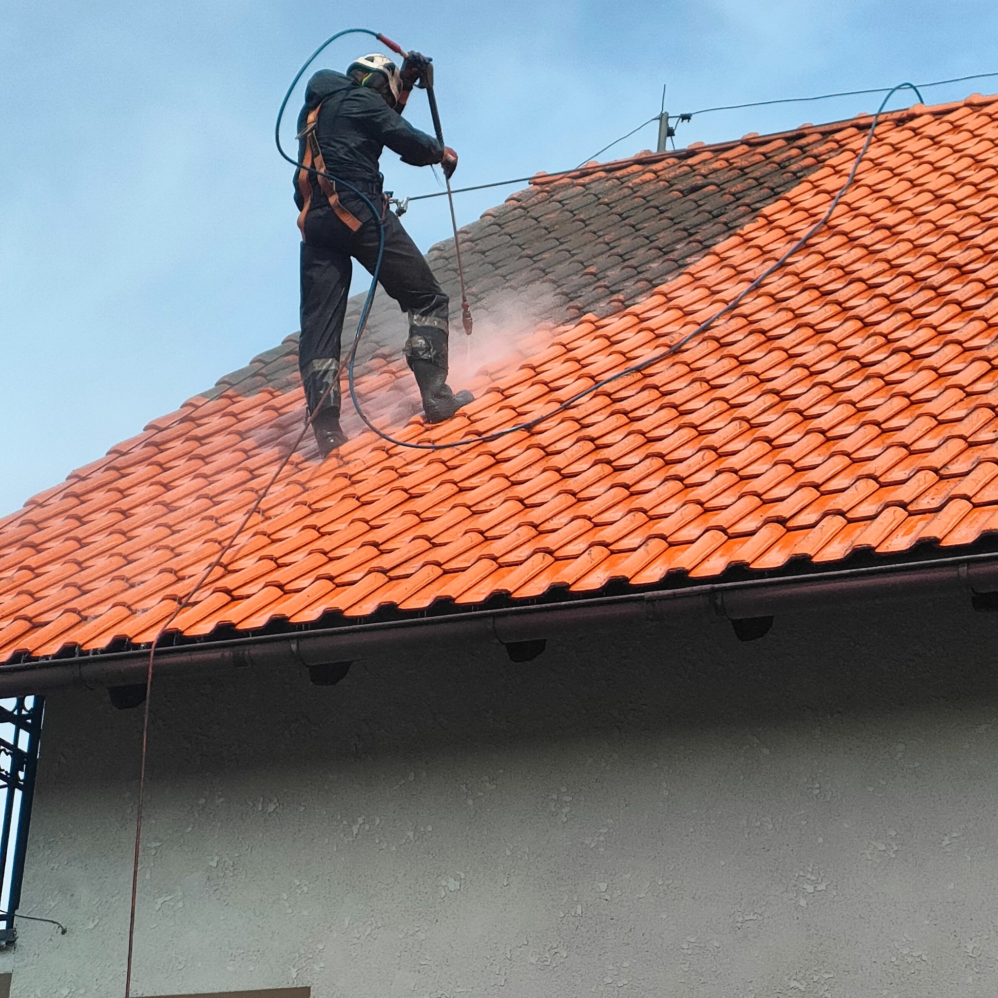 Pracownik w uprzęży myje dachówkę pod ciśnieniem, z wyraźnym efektem czyszczenia - połowa dachu umyta, druga pokryta brudem. Widoczny budynek i niebo.