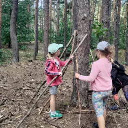 Dwie dziewczynki w lesie budują szałas z gałęzi opierając je o pień drzewa. Jedna z nich ma zielone buty New Balance.