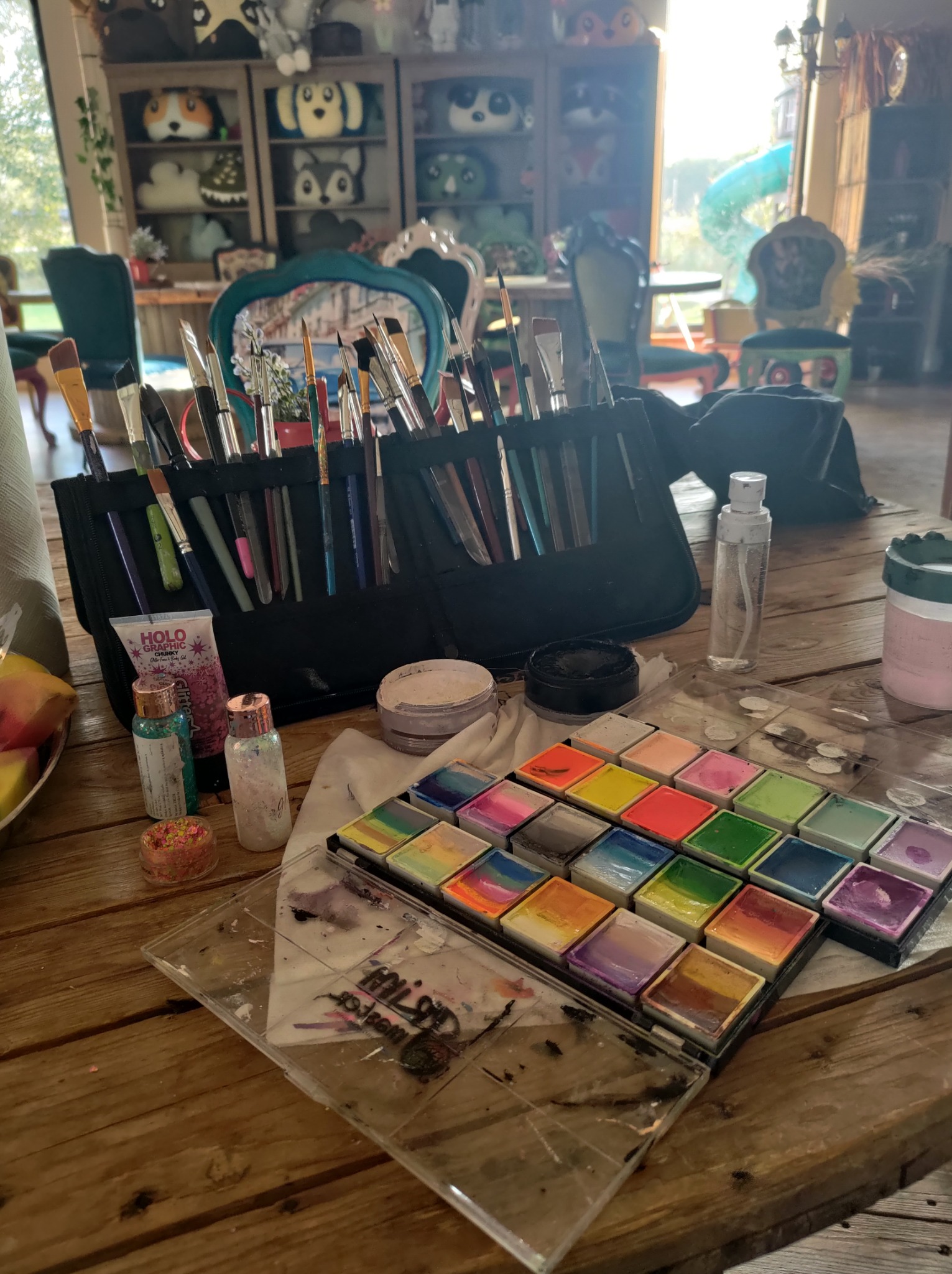 Paleta farb do malowania twarzy, pędzle w etui, brokaty i inne akcesoria na drewnianym stole, w tle pokój z zabawkami i krzesłami.