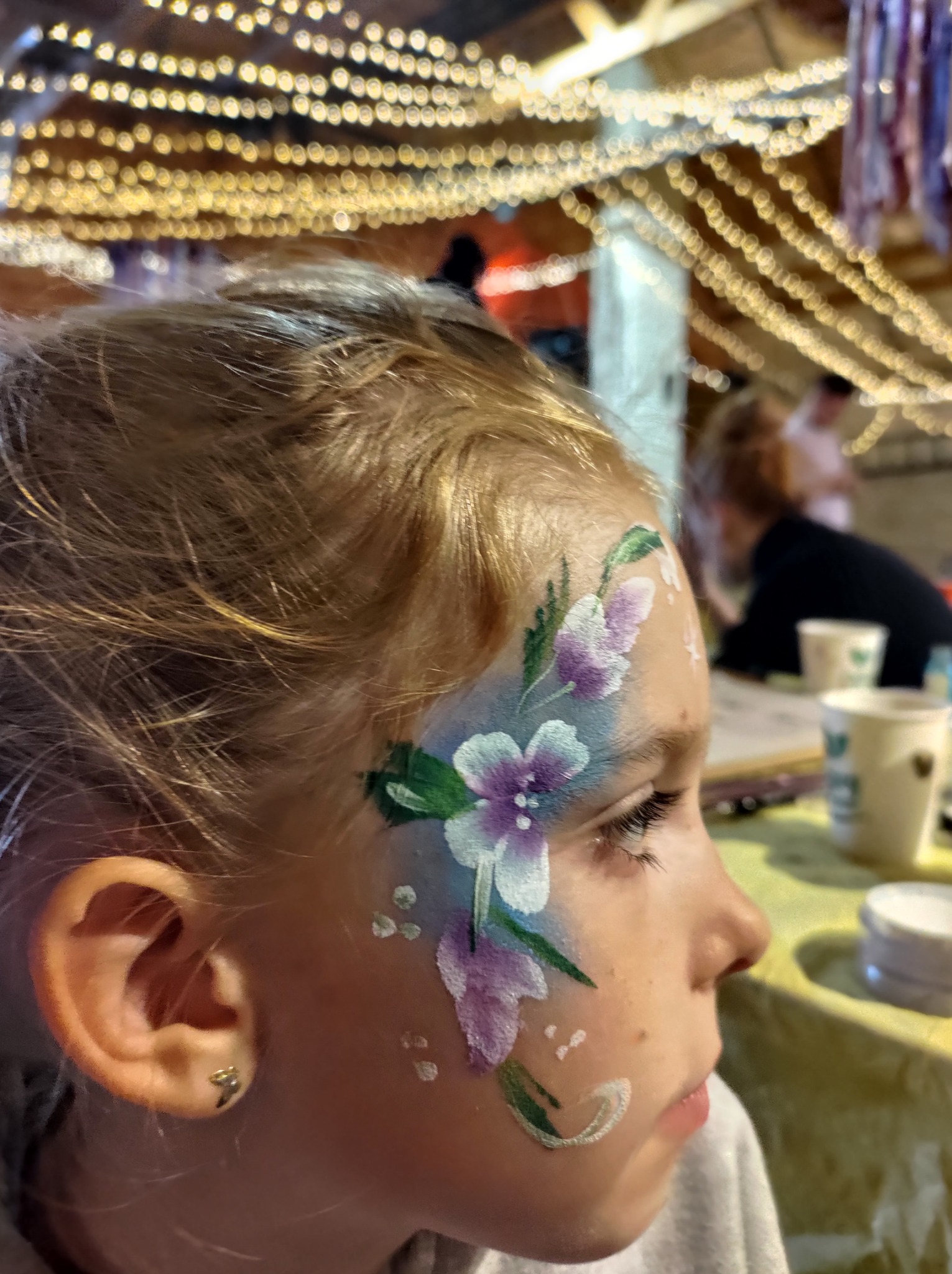 Profil dziewczynki z kwiatowym malunkiem na twarzy, wykonanym farbami w odcieniach fioletu, bieli i zieleni; w tle dekoracje świetlne.