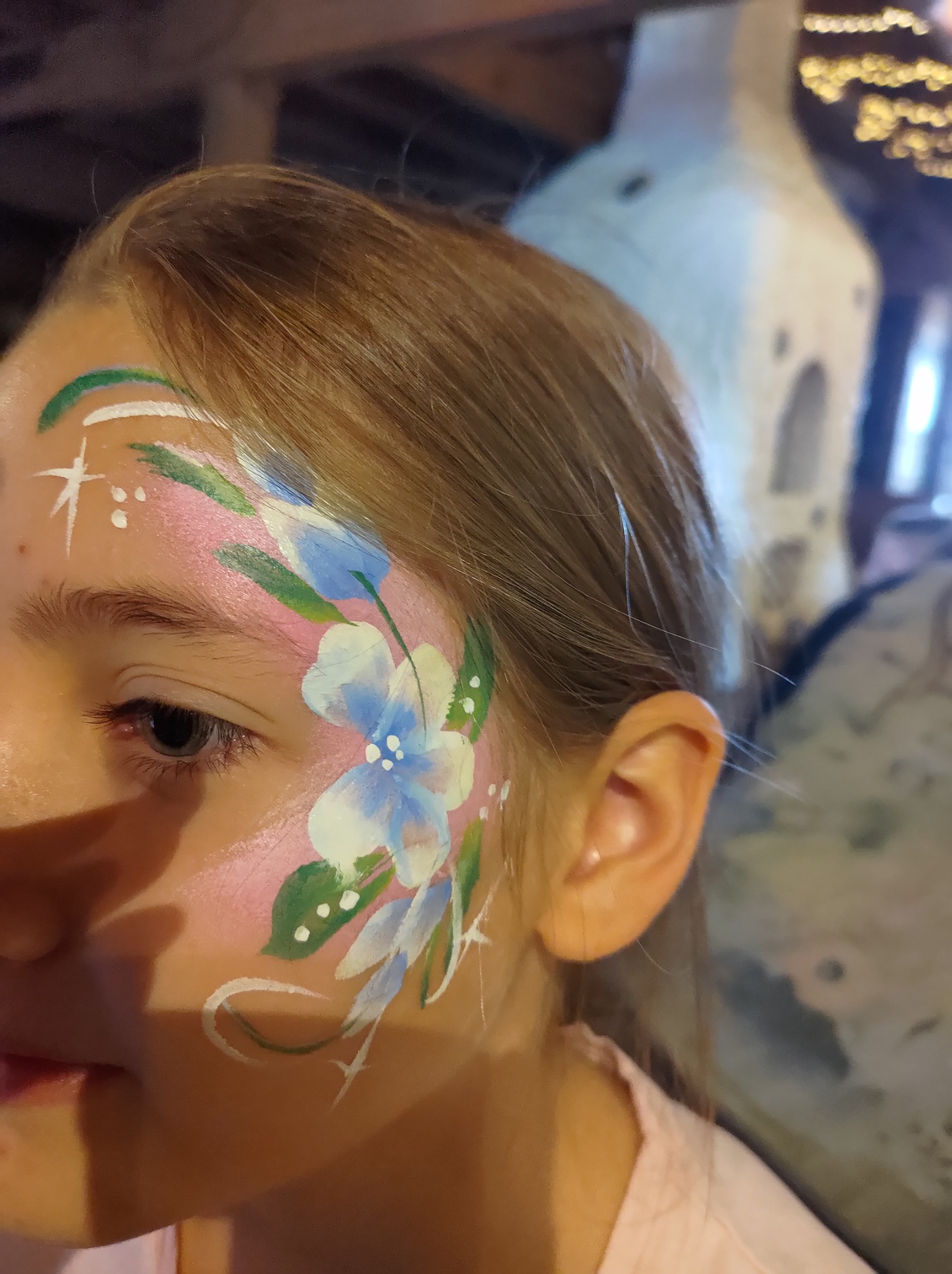 Dziewczynka z pomalowaną twarzą w kwiatowy wzór, z przewagą błękitu i bieli, na tle rozmytego otoczenia.