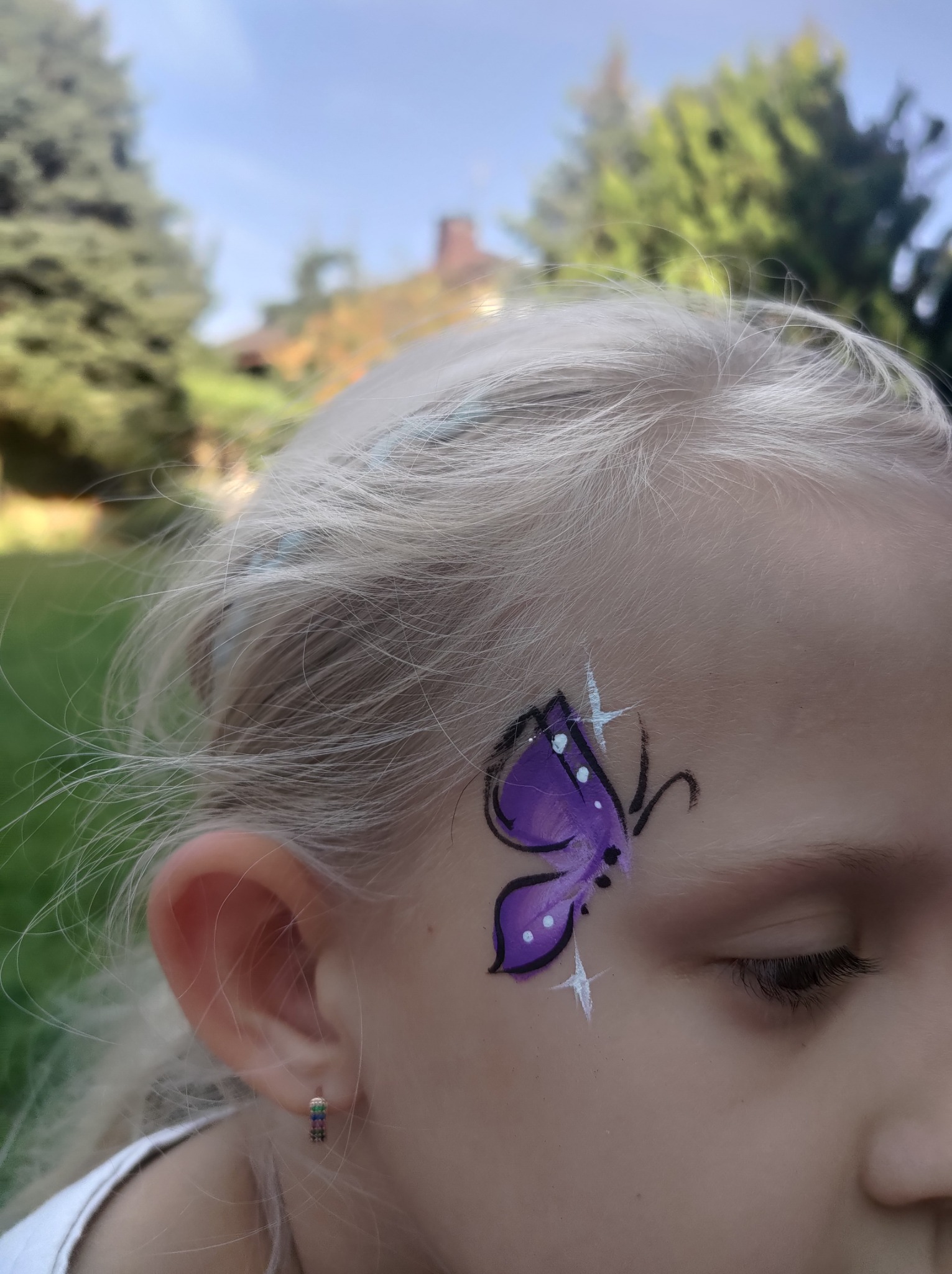 Dziewczynka z pomalowaną na twarzy fioletową motylem z białymi akcentami, widoczne srebrne kolczyki, w tle zieleń drzew i fragment budynku.