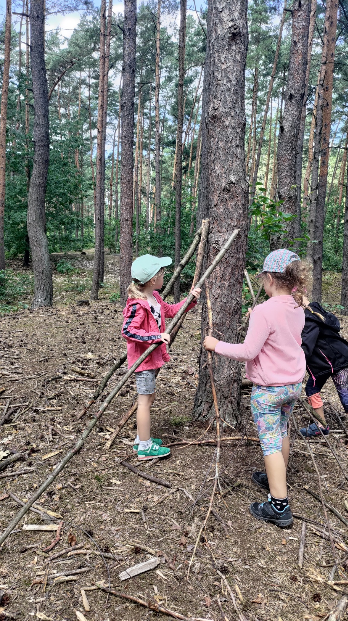 Dwie dziewczynki w lesie budują szałas z gałęzi opierając je o pień drzewa. Jedna z nich ma zielone buty New Balance.
