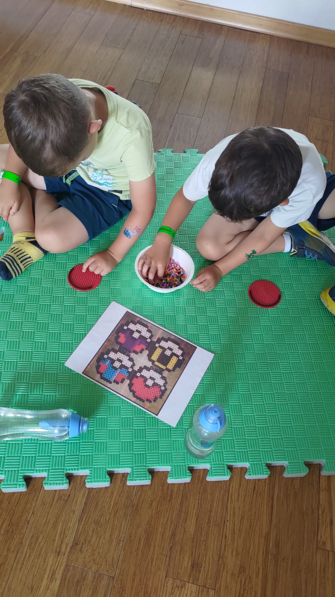 Dwóch chłopców siedzi na zielonej macie i tworzy obrazki z kolorowych koralików, używając podkładek z wypustkami i wzoru na kartce.
