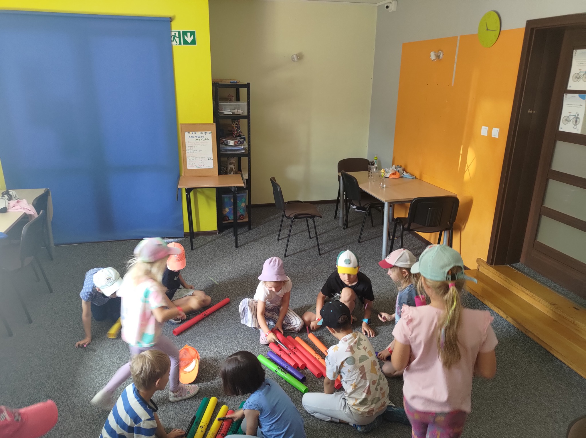 Grupa dzieci siedzi na podłodze w sali o kolorowych ścianach i bawi się kolorowymi rurami bum bum.