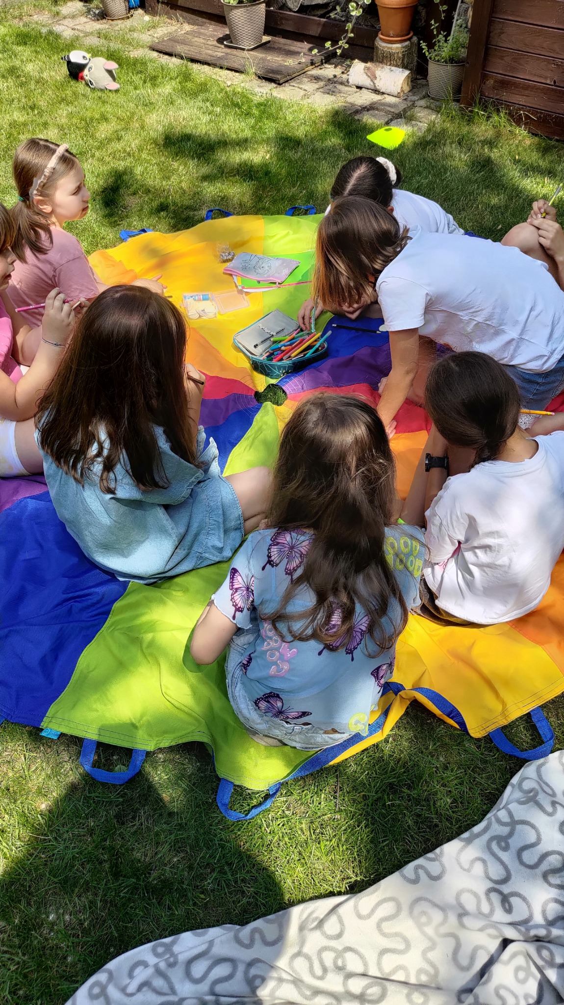 Grupa dzieci siedzi na trawie wokół kolorowej chusty animacyjnej, rysując kredkami i pisakami.