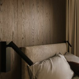 ProHouse - Damian Wąsik - Drewniana ściana za wezgłowiem łóżka z lampką nocną. Minimalistyczny design sypialni z tekstyliami w stonowanych barwach i zasłonami w tle.