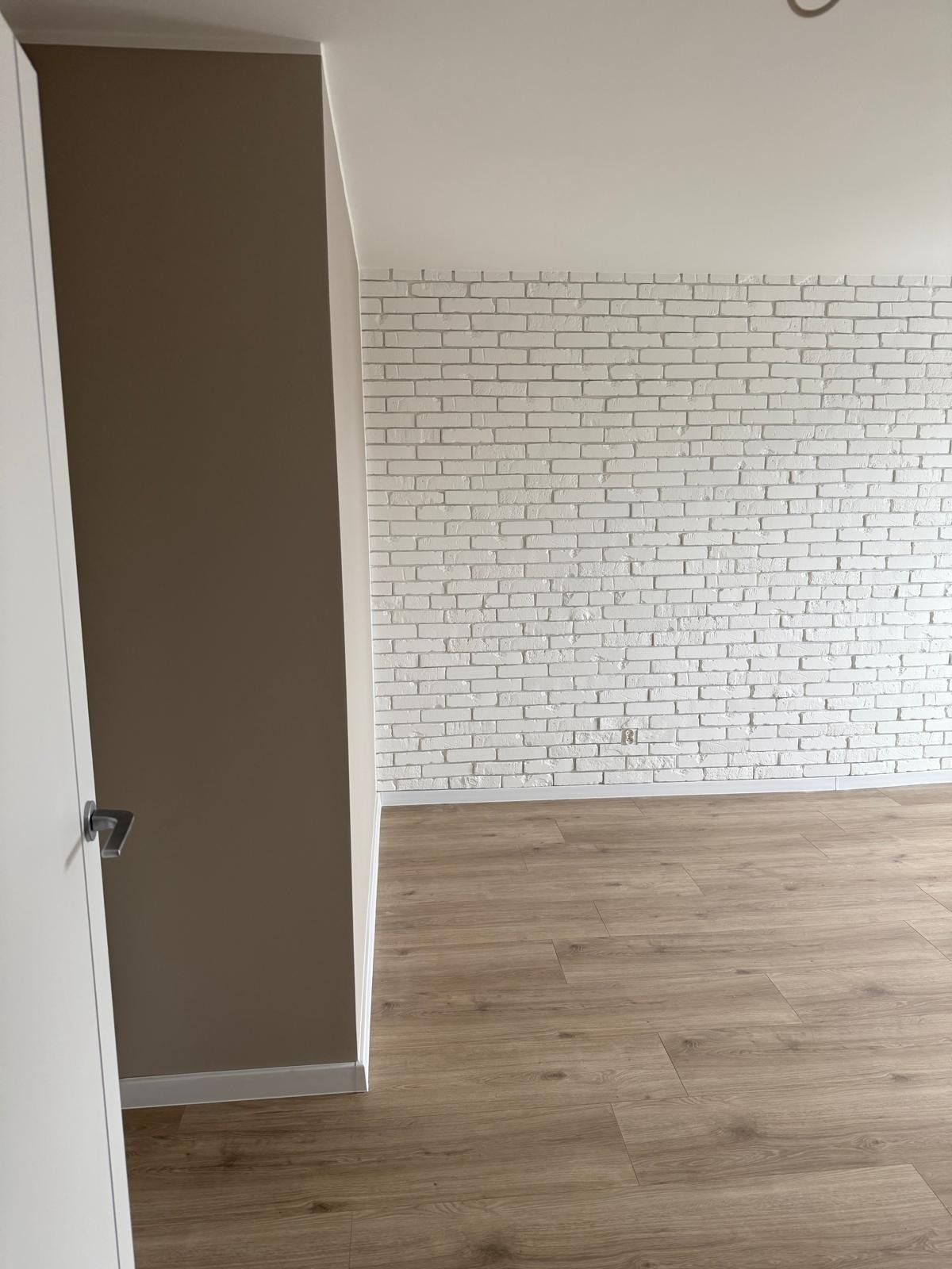 Pomalowane na biało cegły na ścianie, widoczne panele podłogowe, framuga drzwi w kolorze taupe i białe drzwi z klamką.