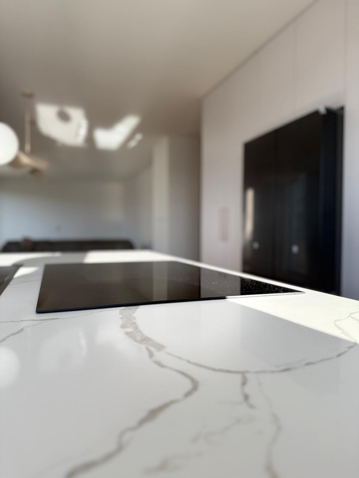 Błyszcząca płyta indukcyjna wbudowana w biały blat z szarymi żyłkami, na tle minimalistycznej kuchni z czarną zabudową.