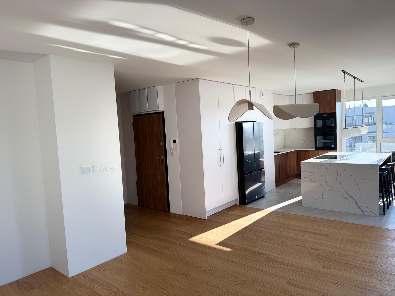 Jasne, przestronne wnętrze mieszkania po remoncie: drewniana podłoga, białe ściany, zabudowa kuchenna z wyspą i nowoczesne lampy wiszące.