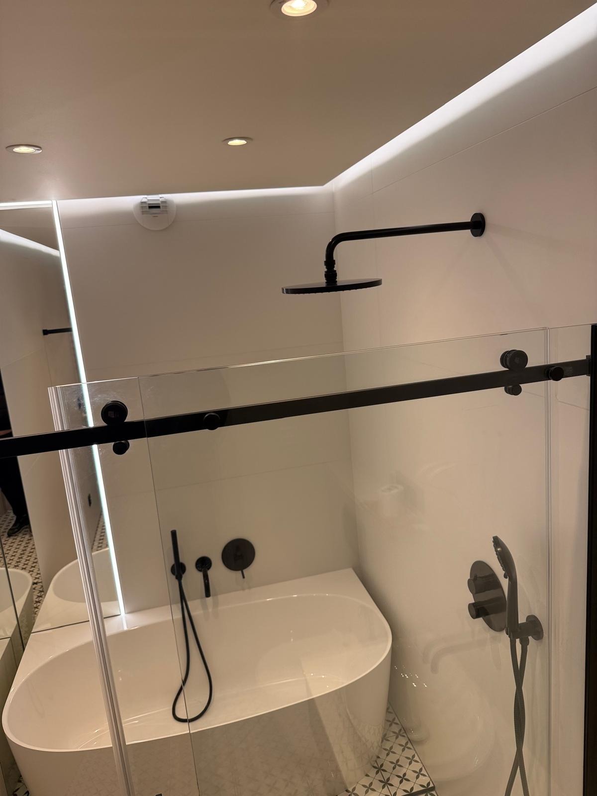 Nowoczesna łazienka z białą wanną, czarną armaturą i szklaną kabiną prysznicową z przesuwnymi drzwiami na czarnych szynach.