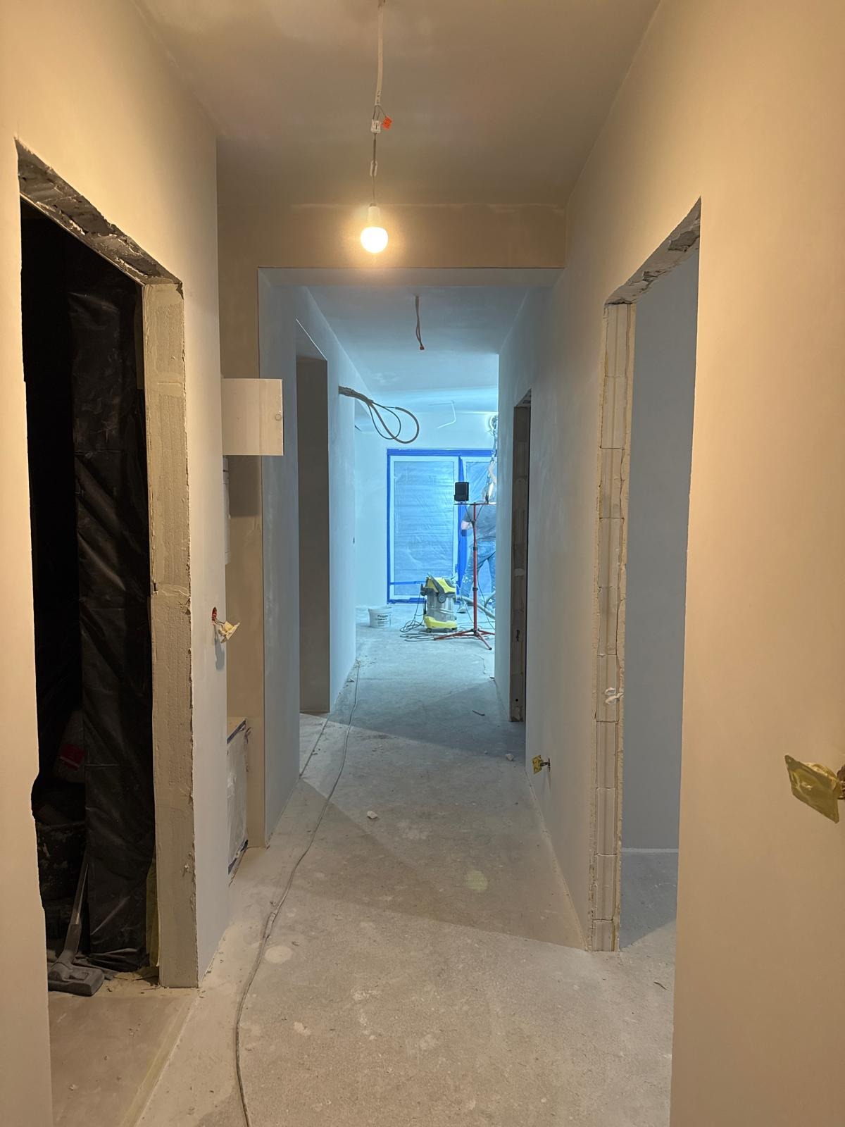Korytarz w trakcie szpachlowania ścian, widoczne przygotowane ościeżnice, kabel wiszący z sufitu i pracownik w oddali z odkurzaczem przemysłowym.