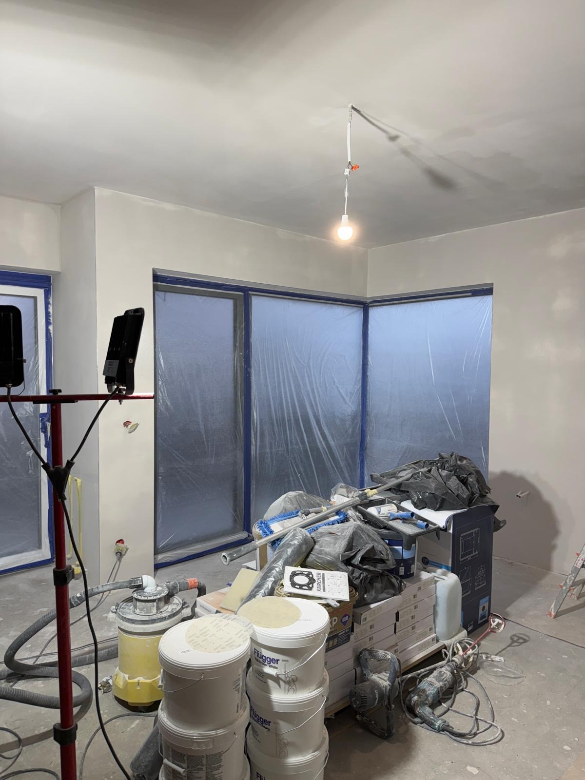 Pokój w trakcie szpachlowania ścian, okna i drzwi zabezpieczone folią, widoczne narzędzia i materiały budowlane, w tym wiadra z masą szpachlową Flűgger i szlifierka do gładzi.