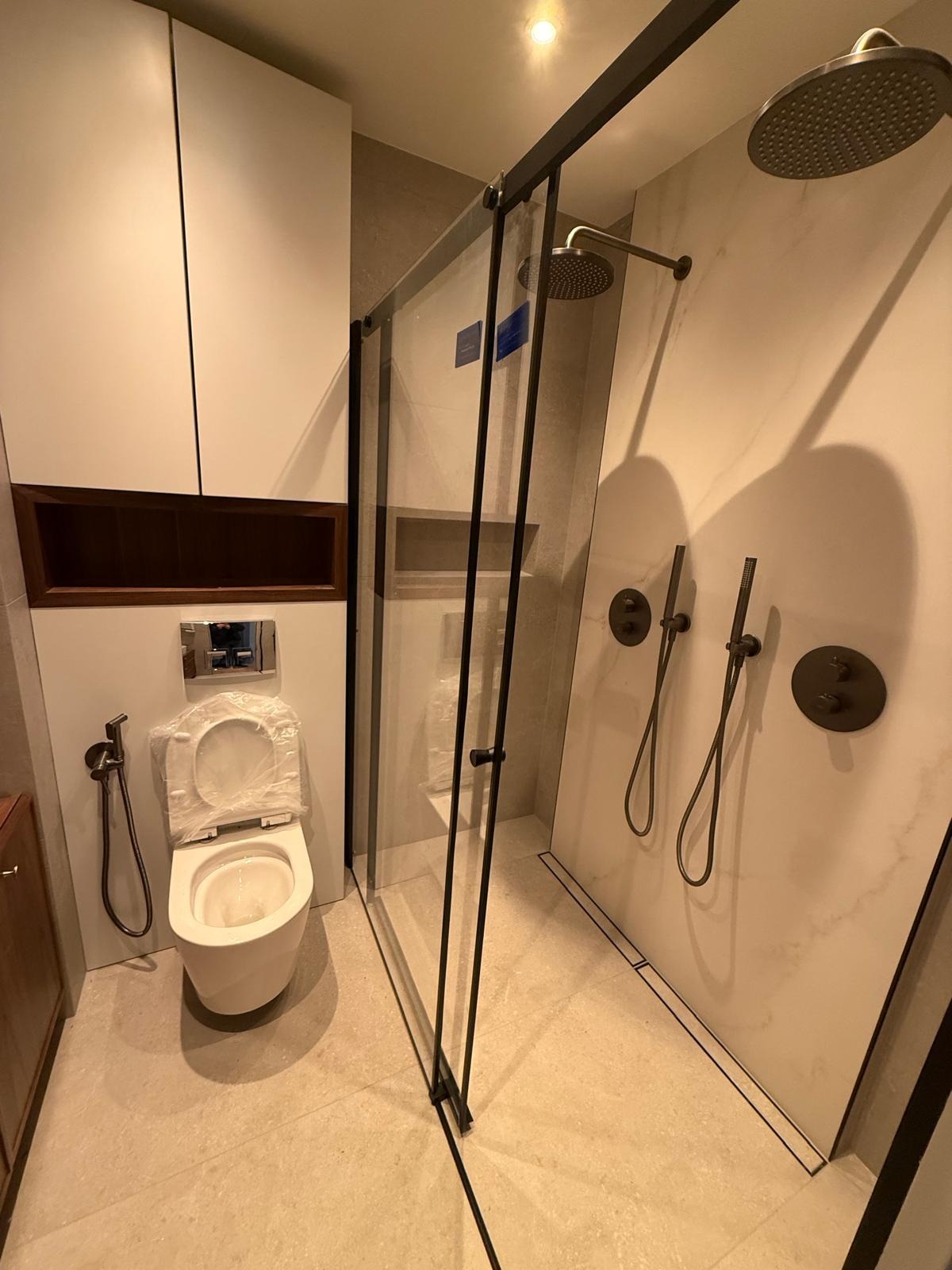 Nowoczesna łazienka z kabiną prysznicową typu walk-in z czarnymi profilami, podwieszaną toaletą z bidetką, zabudową meblową w kolorze białym i brązowym oraz armaturą w kolorze ciemnego brązu.