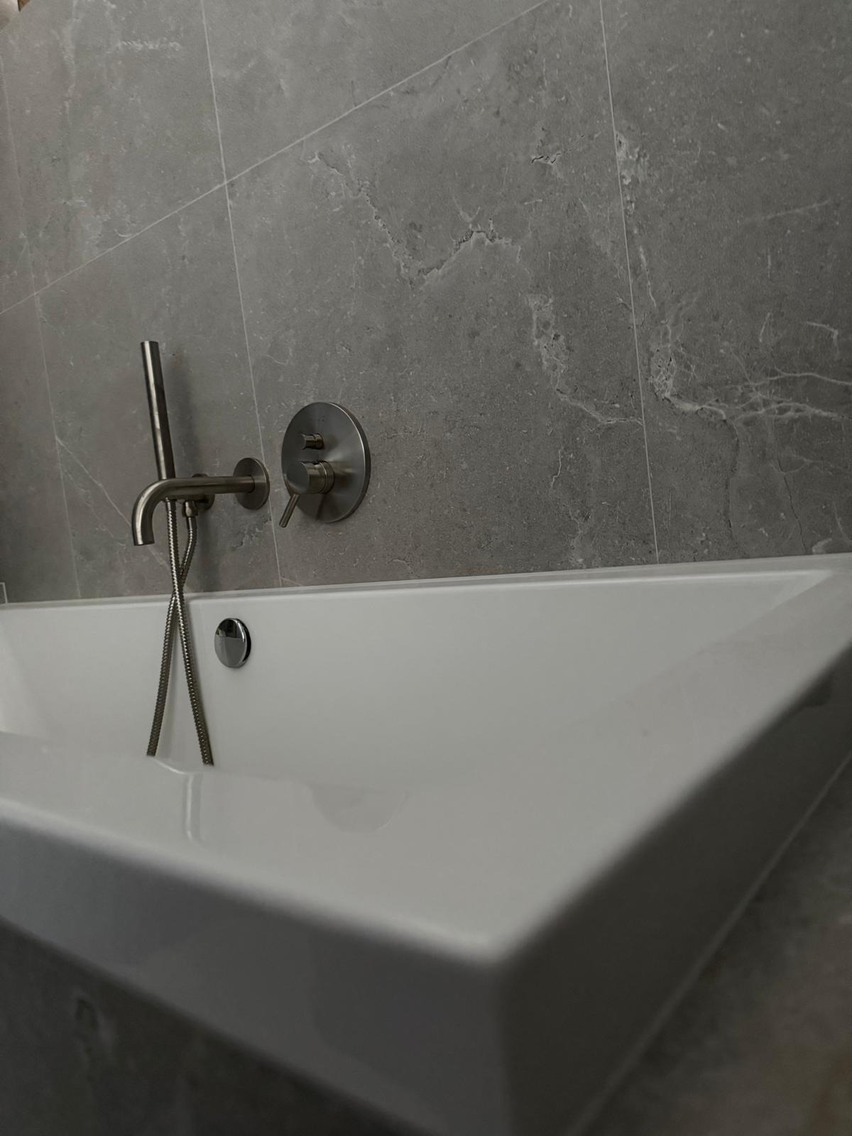 Nowoczesna, biała wanna z chromowaną baterią i słuchawką prysznicową na tle ściany z szarych, marmurkowych płytek.
