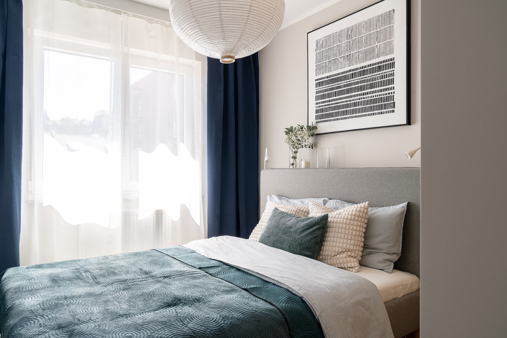 Sypialnia z łóżkiem w odcieniach szarości i turkusu, ozdobiona lampą papierową i grafiką w ramie. Okno z białymi firanami i granatowymi zasłonami.