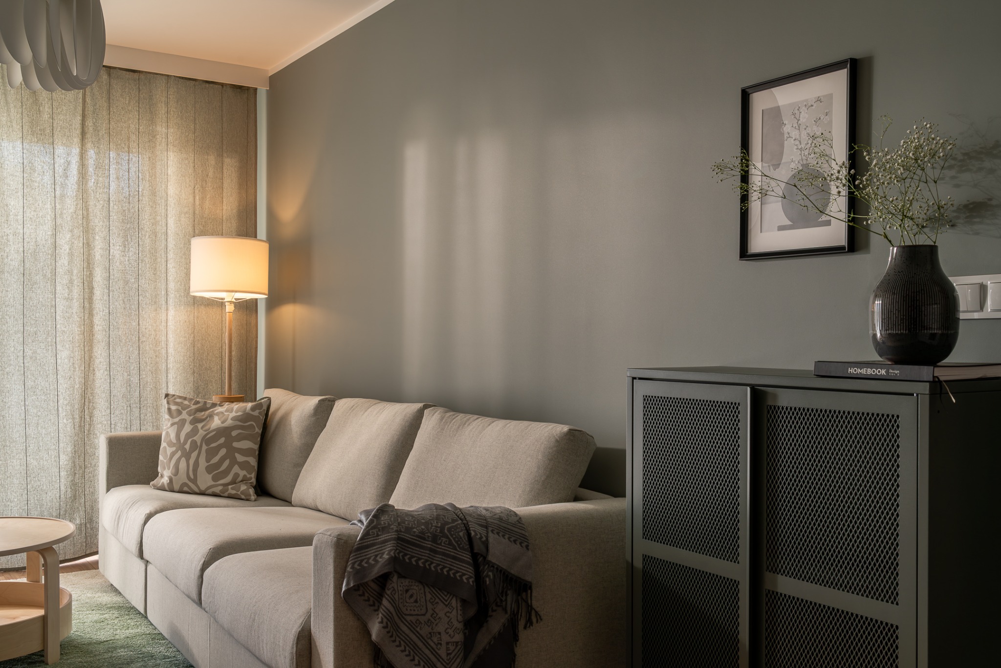 Stylowy salon w odcieniach szarości: sofa z poduszką, stolik, komoda z wazonem i grafika na ścianie. Przytulne, nowoczesne wnętrze z zasłonami.