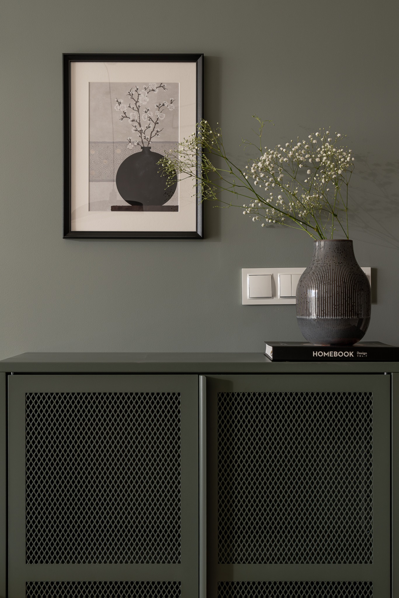 Stylowa komoda z metalową siatką w kolorze khaki, nad nią grafika w czarnej ramie i wazon z gipsówką. Element wykończenia wnętrza w stonowanej kolorystyce.