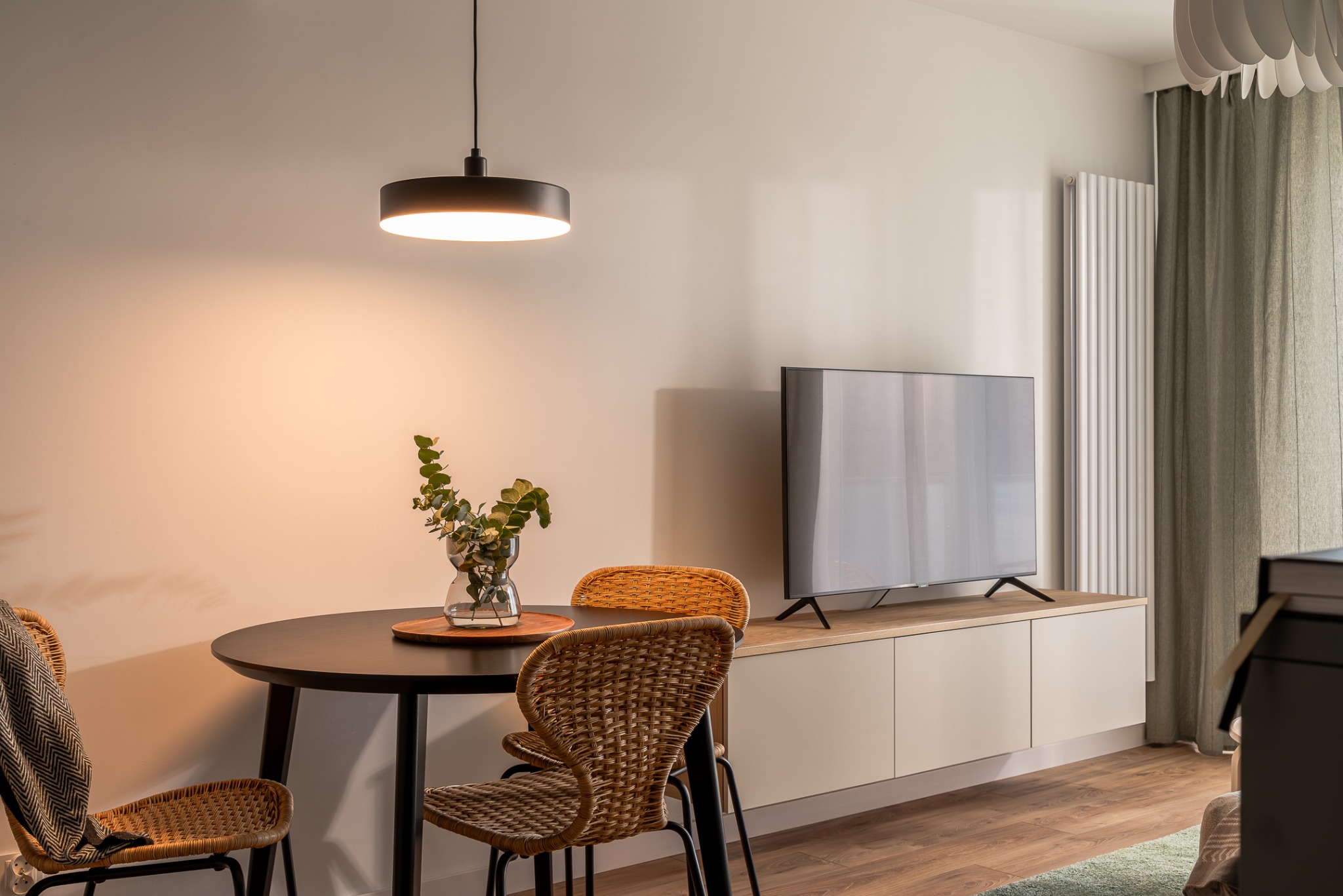 Jasne wnętrze: okrągły stół z bukietem, rattanowe krzesła, szafka RTV z telewizorem. Minimalistyczny design, drewniana podłoga i ciepłe oświetlenie.