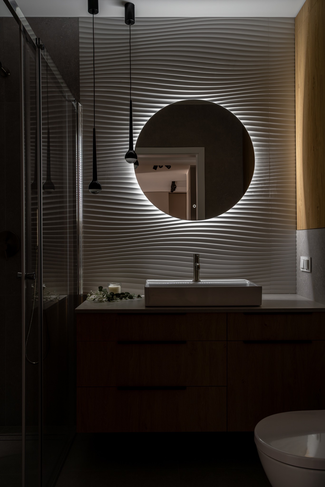 Nowoczesna łazienka z umywalką nablatową, okrągłym lustrem z podświetleniem LED i dekoracyjną ścianą z falistych paneli 3D. Minimalistyczne lampy wiszące.