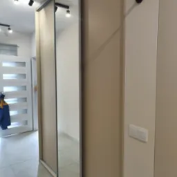 Beżowa szafa z przesuwnymi lustrzanymi drzwiami we wnęce, widoczny fragment sufitu z oświetleniem punktowym oraz drążek do podciągania zamontowany nad szafą.